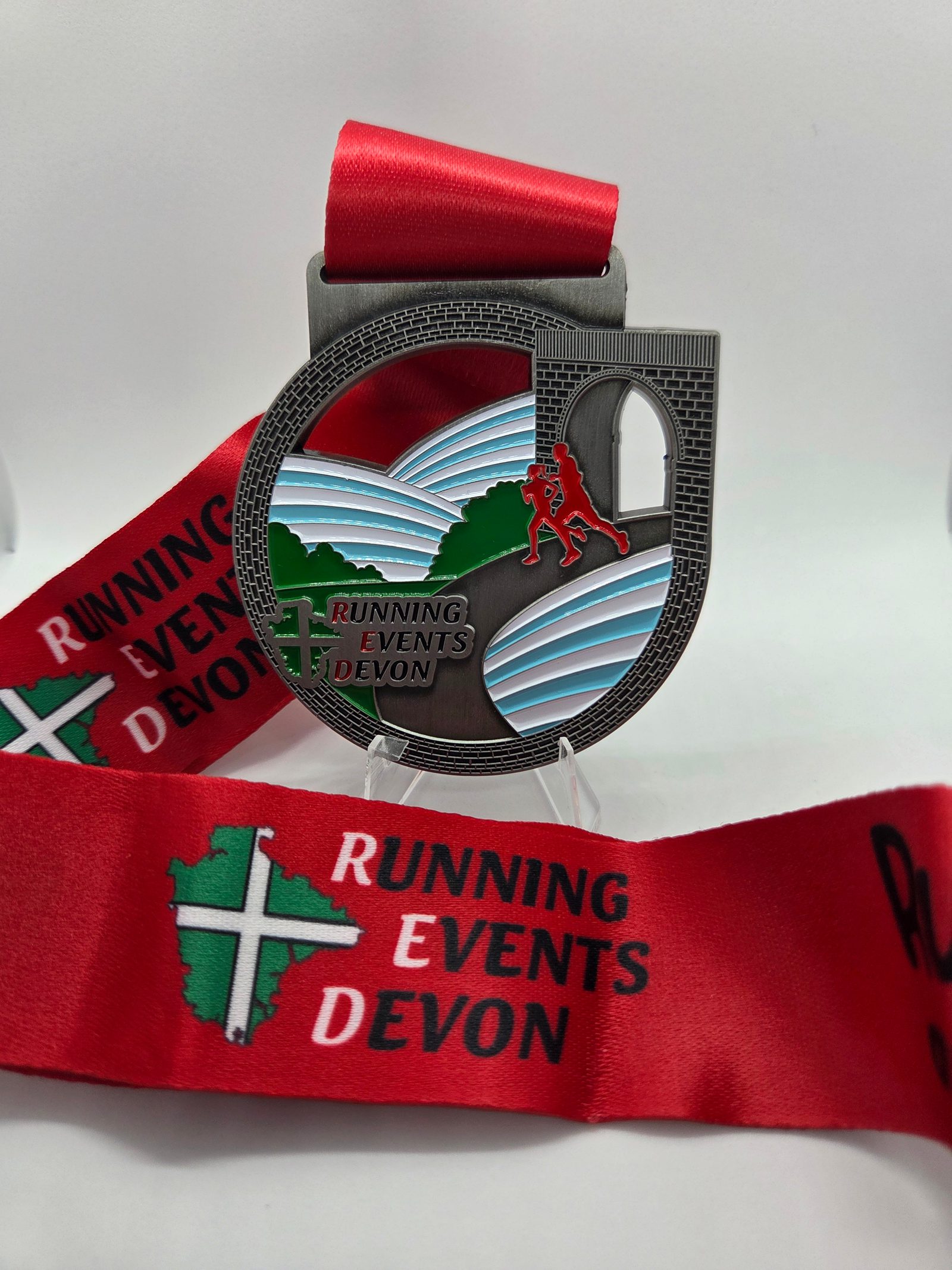 A silver medal with a red ribbon that reads "Running Events Devon." The medal features a runner, a bridge, hills, and the event name with a cross-shaped map of Devon in green.