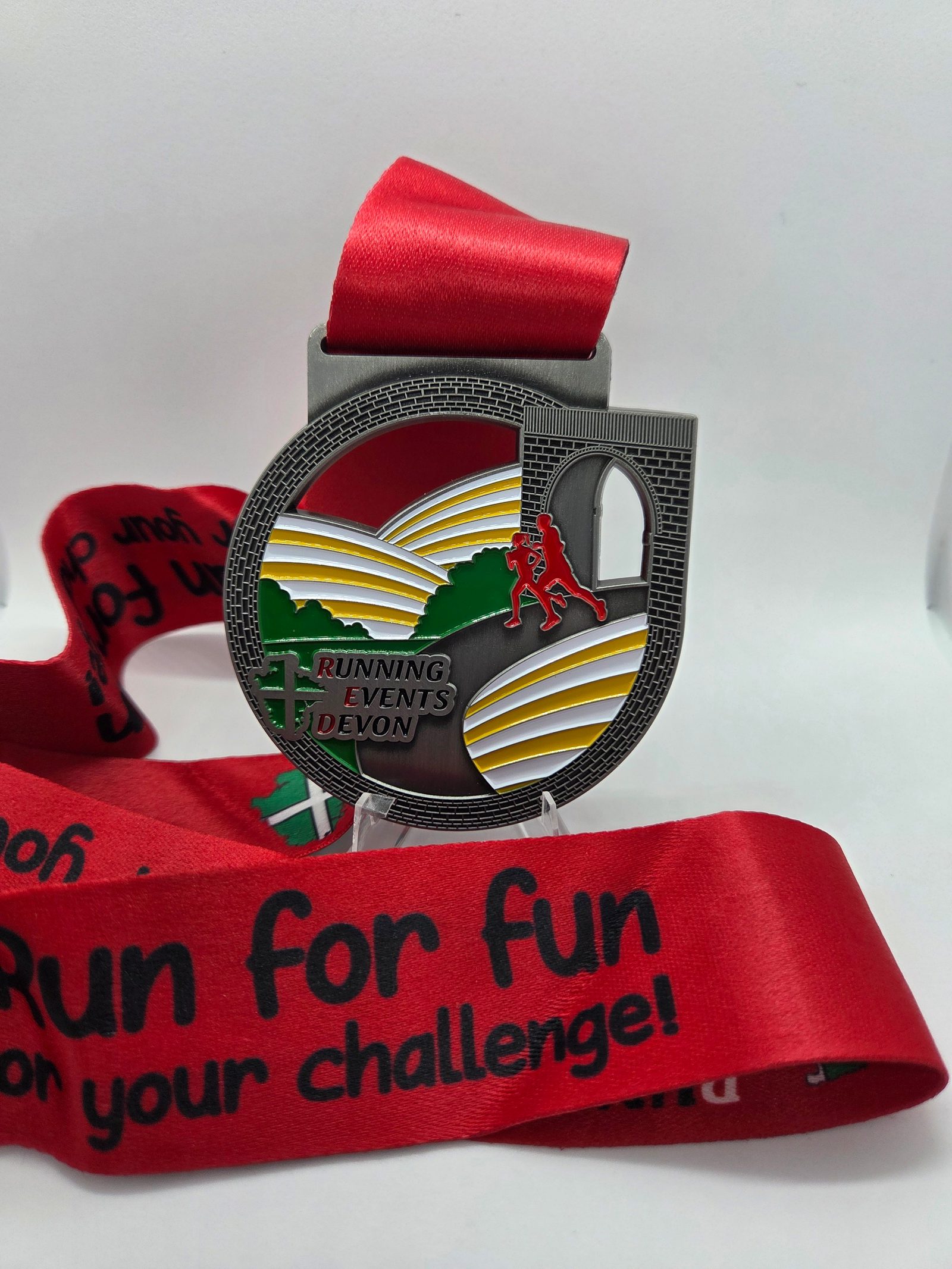 A silver medal with a red ribbon features a landscape, a viaduct, and a red runner, with "Running Events Devon" written on it. The ribbon says, "Run for fun or for your challenge!" in bold black text.