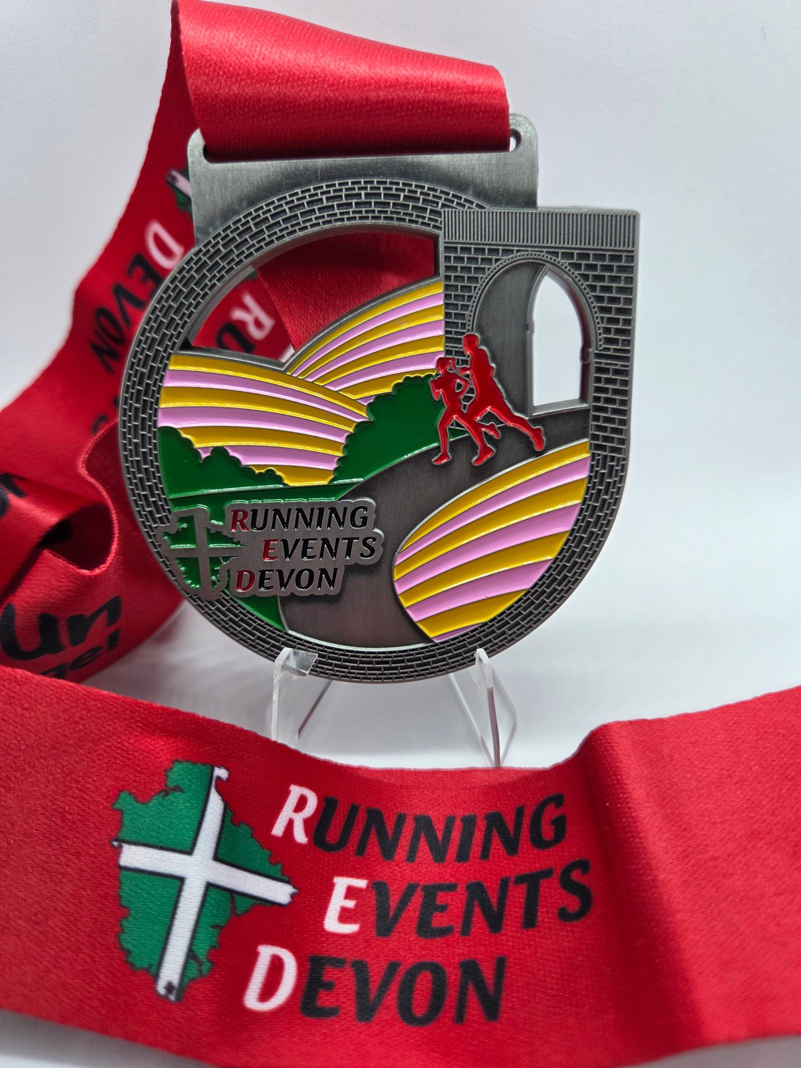 A silver medal with colorful hills, a bridge, and two running figures, labeled "Running Events Devon." The medal hangs on a red ribbon with the same text and a green map graphic.