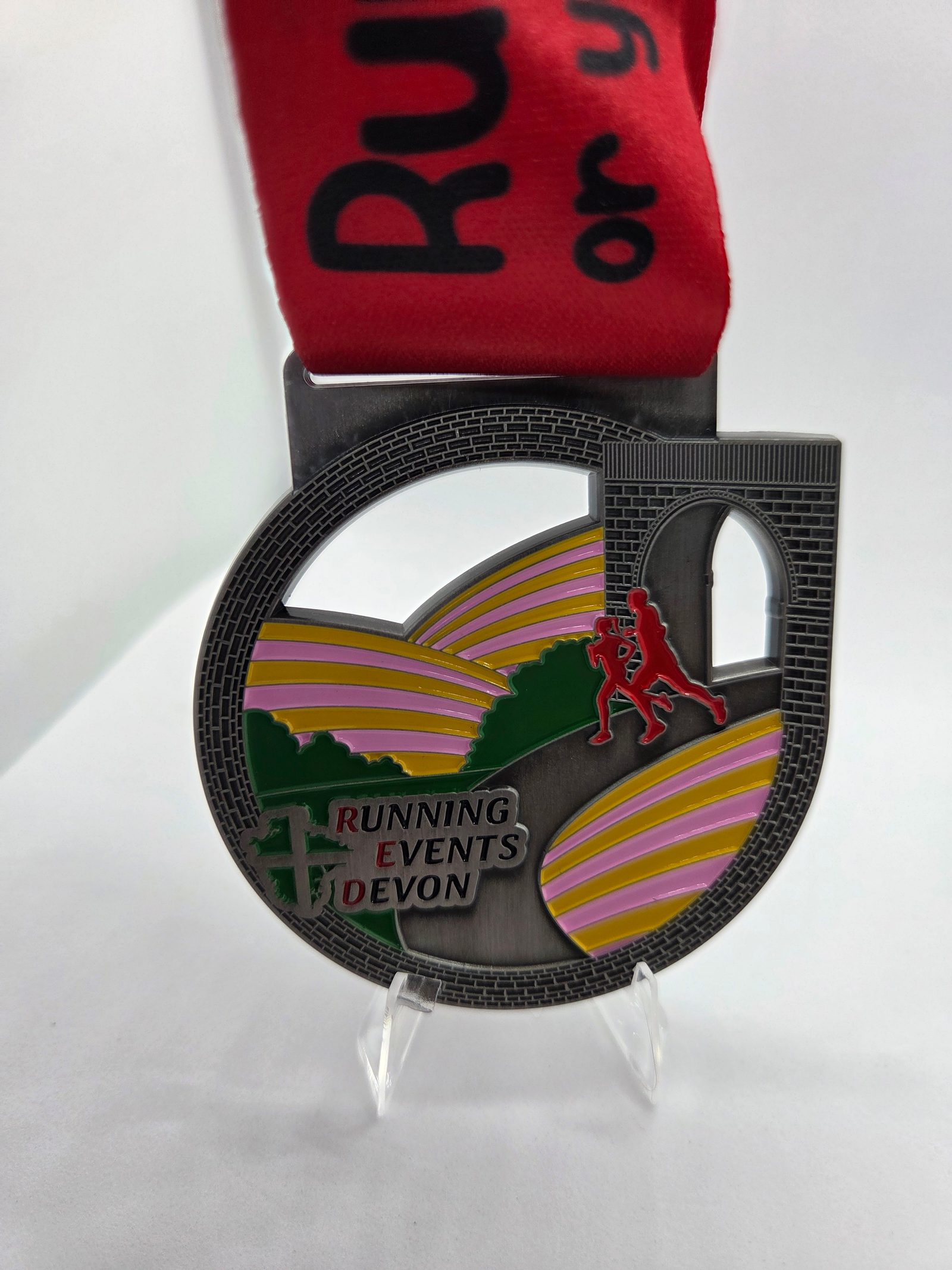 A circular medal with a red ribbon, featuring a runner figure, rolling hills with pink and yellow stripes, an archway, and the text “Running Events Devon.” The medal is displayed on a clear stand.
