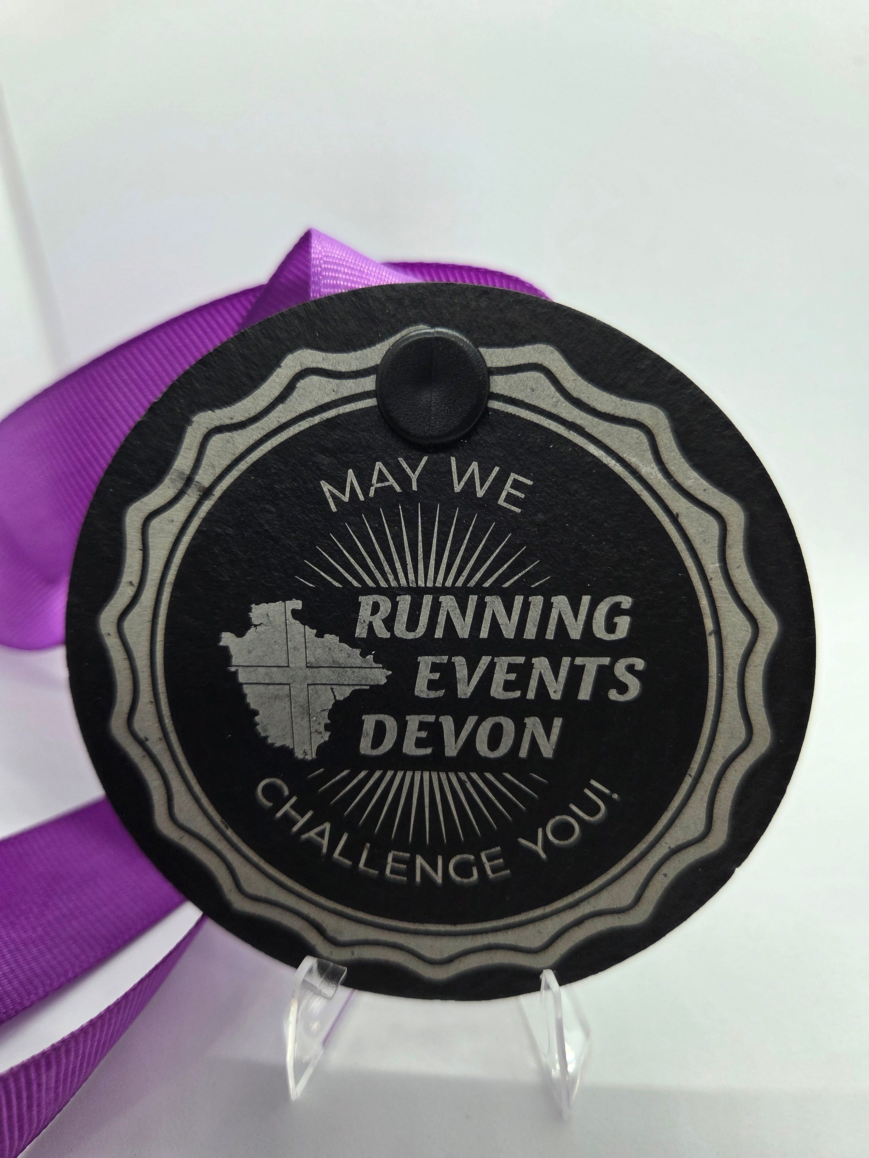 A round black medal with "May We Challenge You! Running Events Devon" written on it, featuring a map outline, and attached to a purple ribbon, displayed on a clear stand against a white background.