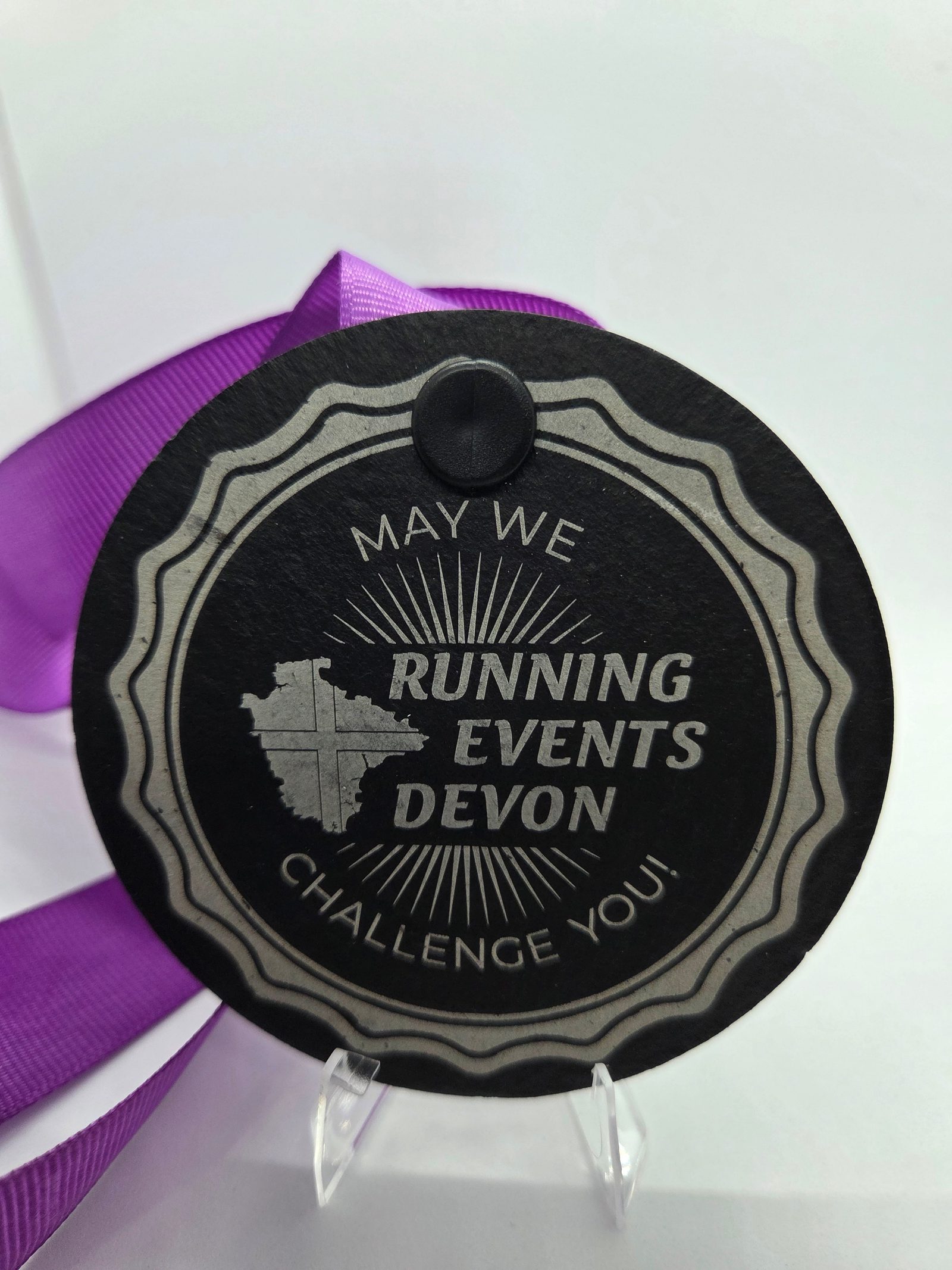 A round black medal with a wavy silver border reads "May We Challenge You! Running Events Devon" with an outline of Devon and a sunburst design, attached to a purple ribbon.