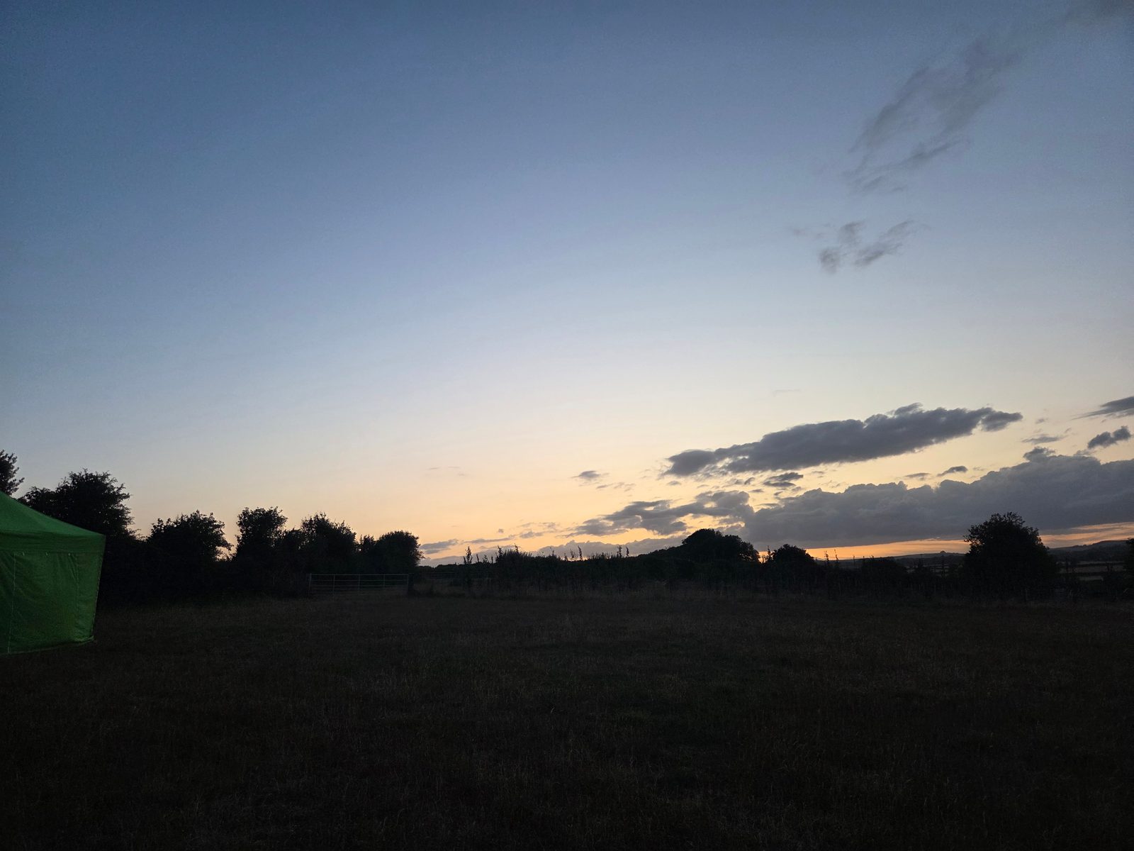 A grassy field at dusk with a silhouette of trees on the horizon, a blue and orange sky with scattered clouds, and a green tent partially visible on the left side.