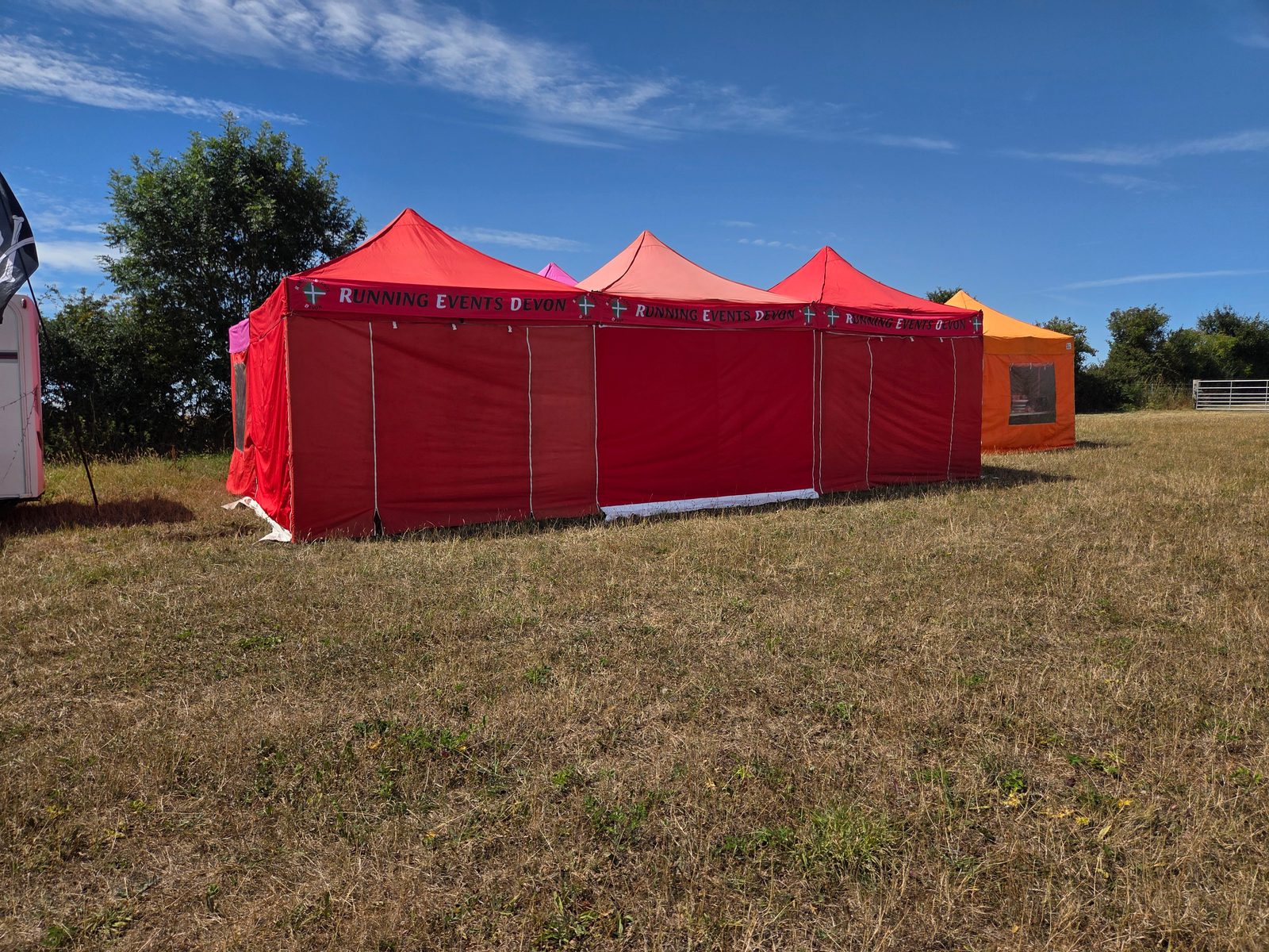 Three connected red event tents labeled "RUNNING EVENTS DEVON" stand on dry grass under a blue sky, with an orange tent and trees in the background.
