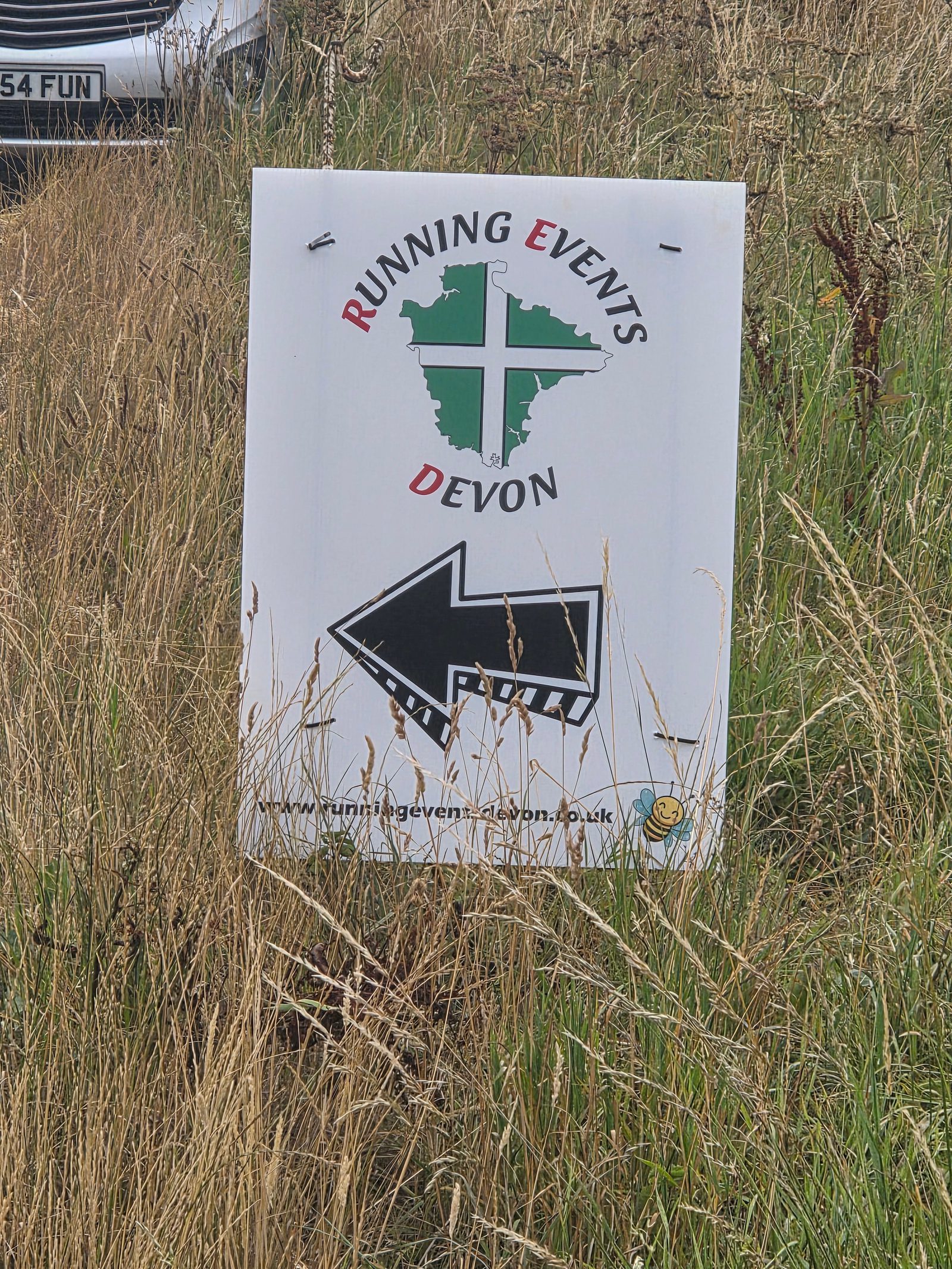 A sign in tall grass reads “Running Events Devon” with a map graphic and black left arrow, directing participants. The website runningeventsdevon.co.uk is printed at the bottom.