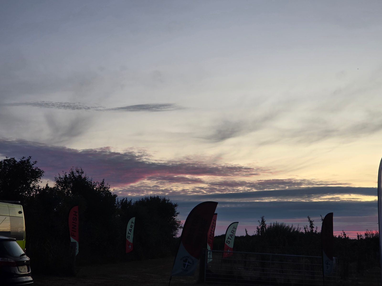A dusky sky with streaks of pink and purple clouds above silhouetted trees. Several flag banners, some labeled "FINISH," stand along a dirt path next to parked vehicles.
