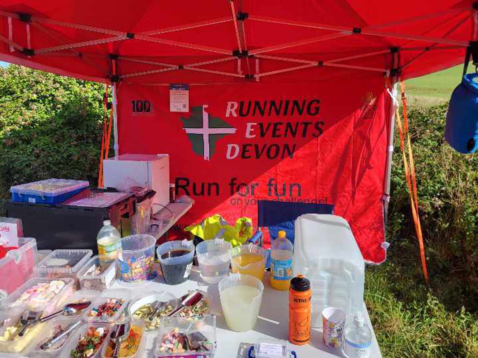 A red tent with a "Running Events Devon" banner is set up beside a green field. The foreground displays a table cluttered with various snacks, drinks, and running supplies. Mixed beverages and food items are arranged in plastic containers.