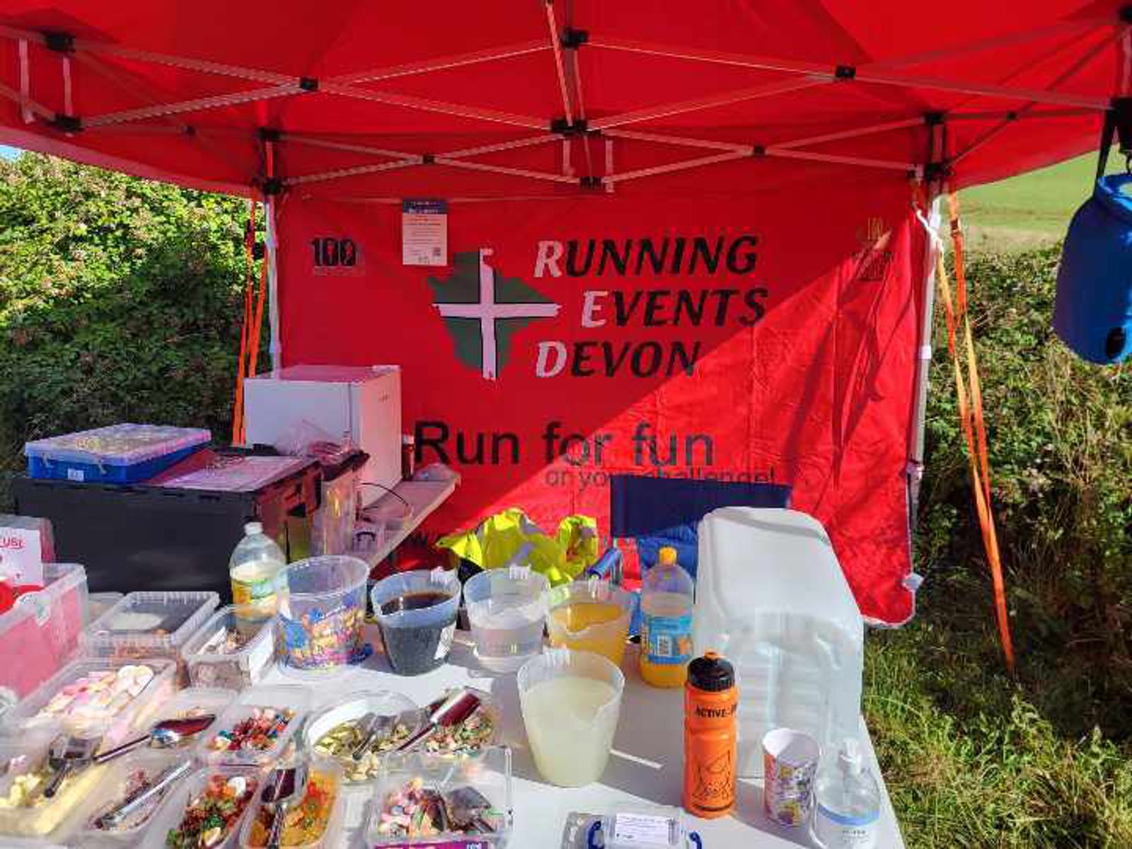 A red tent at an outdoor event hosted by Running Events Devon. The table inside has various food items, drinks, and supplies. The backdrop displays the event's logo and slogan "Run for fun." The surroundings include greenery under a clear sky.