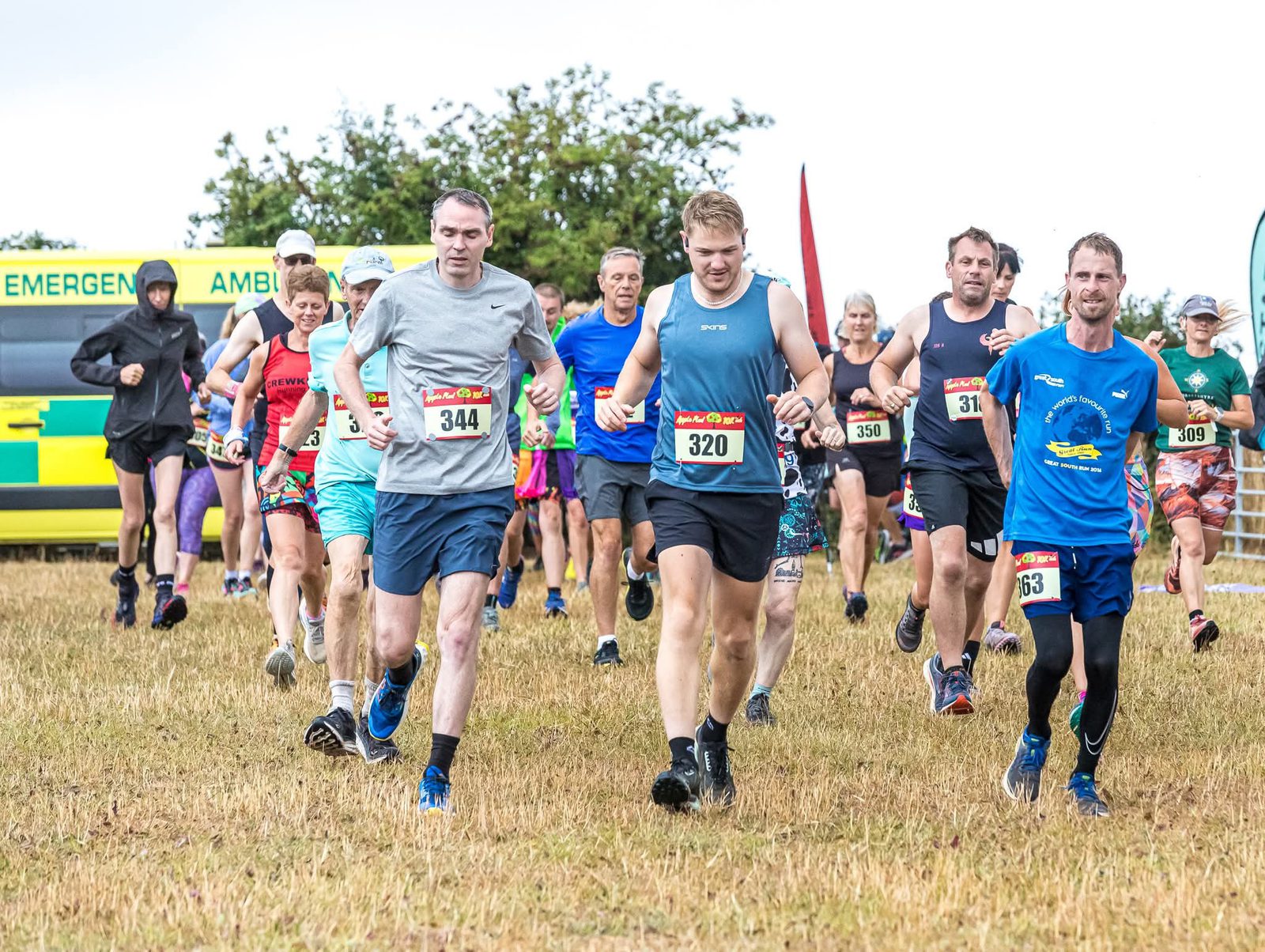 A group of runners in athletic gear participate in a race on a grassy field, with race numbers pinned to their shirts and an emergency ambulance visible in the background.