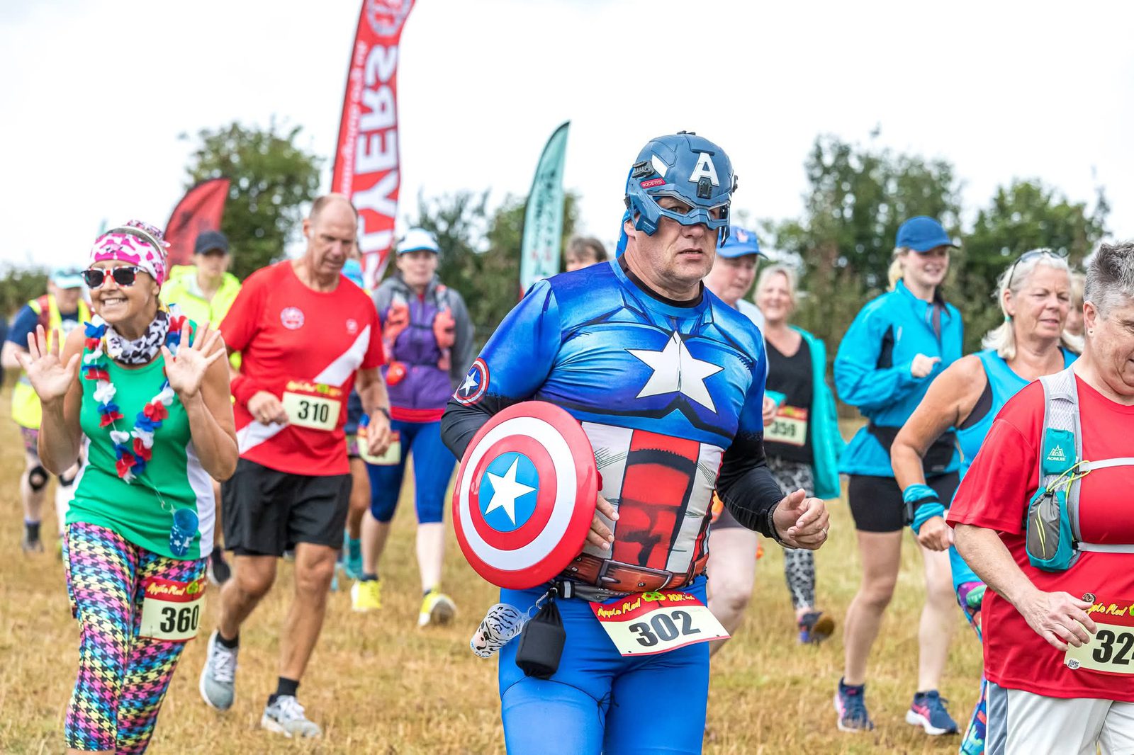 A group of runners in colorful outfits, including a man dressed as Captain America with a shield and mask, participate in an outdoor race on a grassy field. Other runners wear bright costumes and numbered bibs.