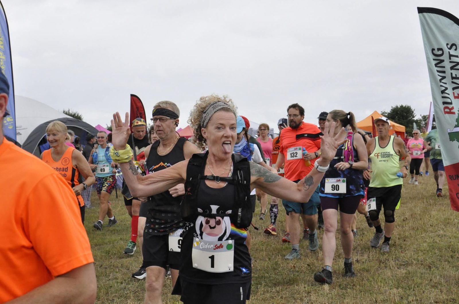 A group of runners participate in a race outdoors. A woman in front with curly hair, wearing bib number 1, sticks out her tongue and raises her hands energetically, while other runners follow behind her.