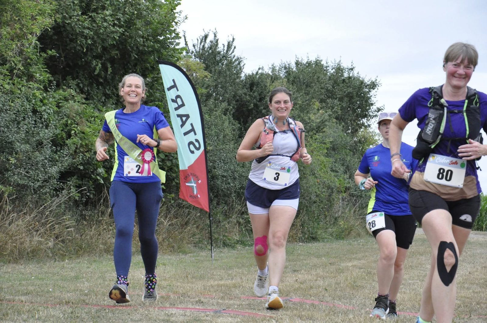 Four women are running in a race, smiling as they pass a "START" flag on a grassy path with trees in the background. Each wears a bib number and athletic gear, appearing energetic and happy.