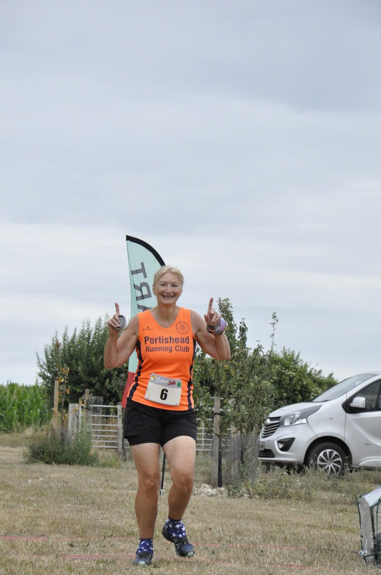 A smiling runner in an orange "Portishead Running Club" shirt and black shorts flashes peace signs while running outdoors, with trees, a car, and a club flag in the background.