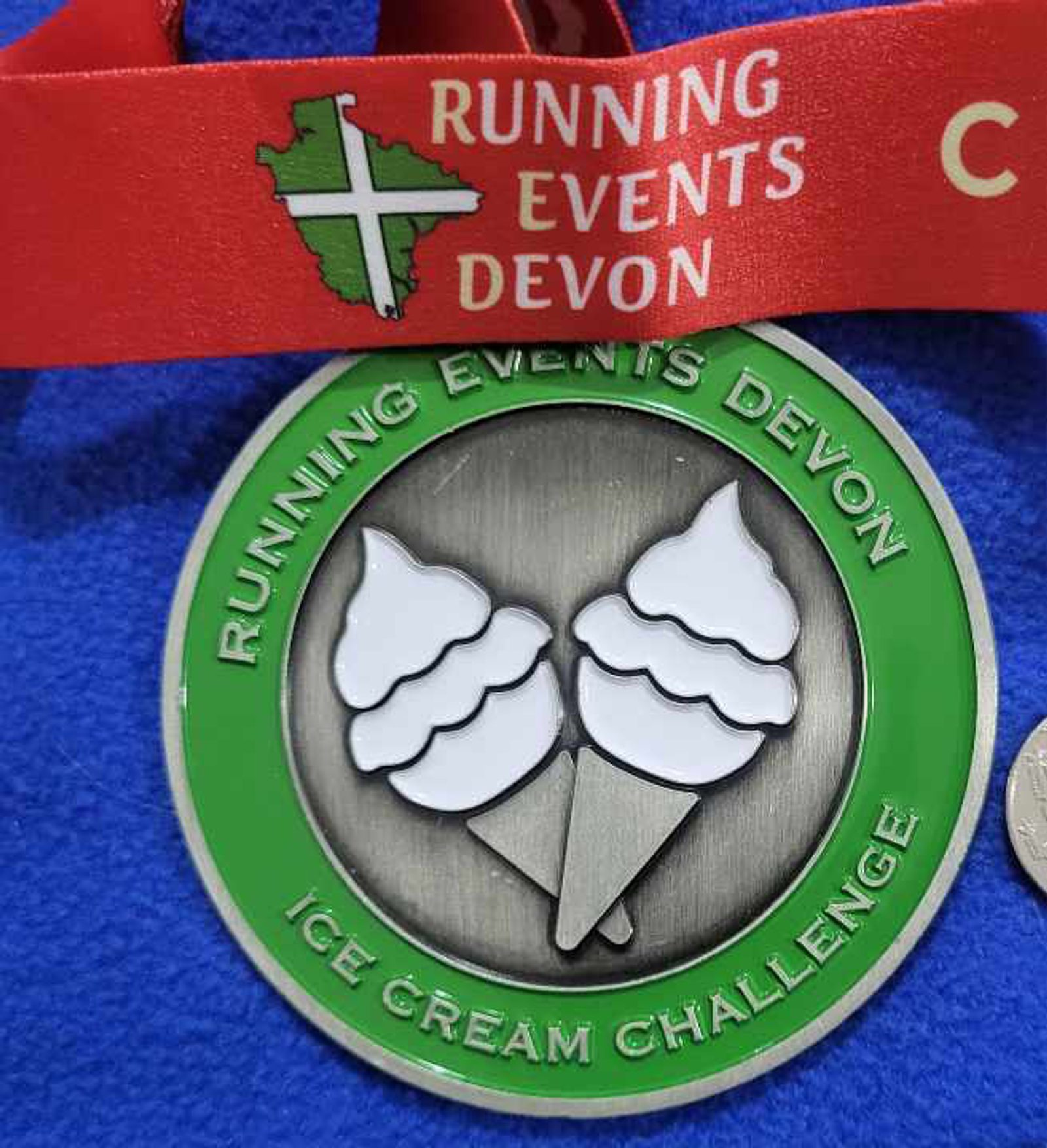 A circular medal with a green border and a silver center featuring two ice cream cones. The text around the center reads "Running Events Devon" on the top and "Ice Cream Challenge" on the bottom. The medal is attached to a red ribbon.
