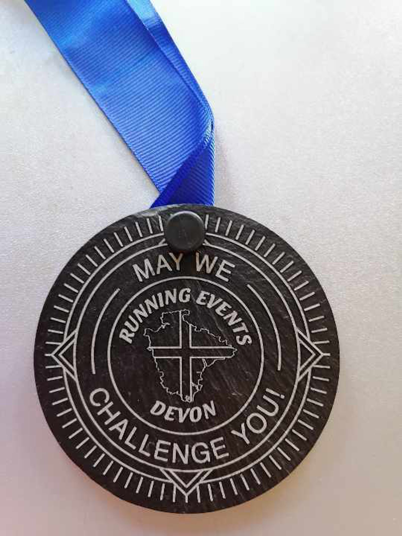 A round, black medal with a blue ribbon is inscribed with "May We Challenge You! Running Events Devon" and features a map of Africa with a cross.
