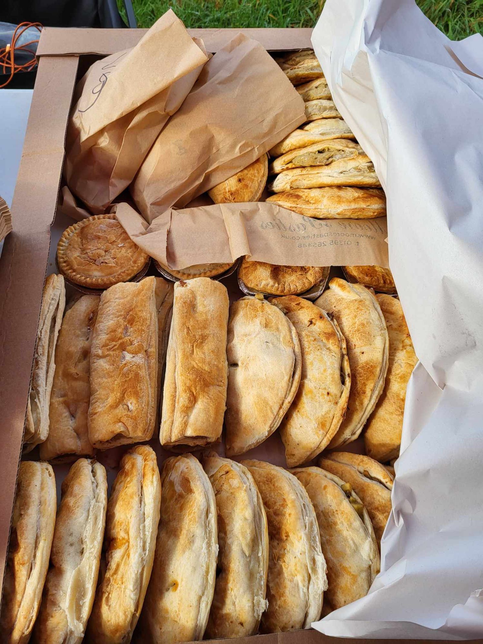 A large cardboard box is filled with various baked goods, including numerous pasties, pies, and some items wrapped in brown paper. The assortment appears freshly baked with golden brown crusts, neatly arranged, and ready to be served.