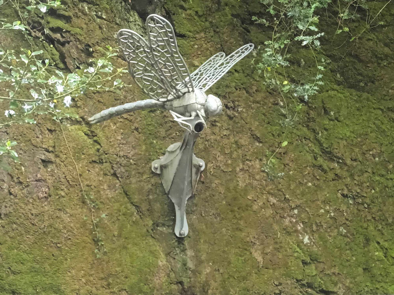 A metallic dragonfly sculpture is attached to a moss-covered rock face. It has intricately detailed wings and a body resembling mechanical parts, blending nature and industrial art. Greenery surrounds the sculpture, enhancing its integration into the natural environment.