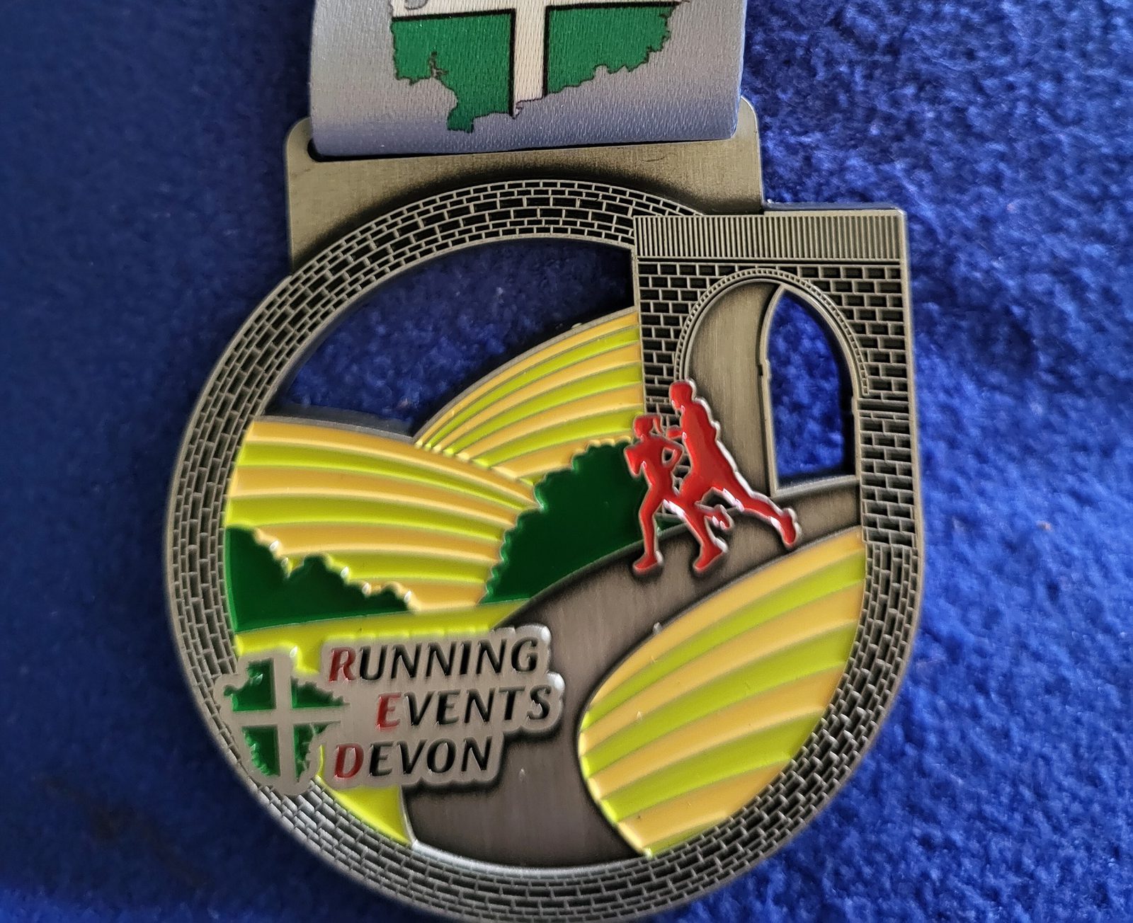 A medal featuring two red silhouettes of runners crossing a bridge above green and yellow fields. The top of the medal has a map outline with a white cross on a green background. Below the runners, text reads "RUNNING EVENTS DEVON." The background is blue fabric.