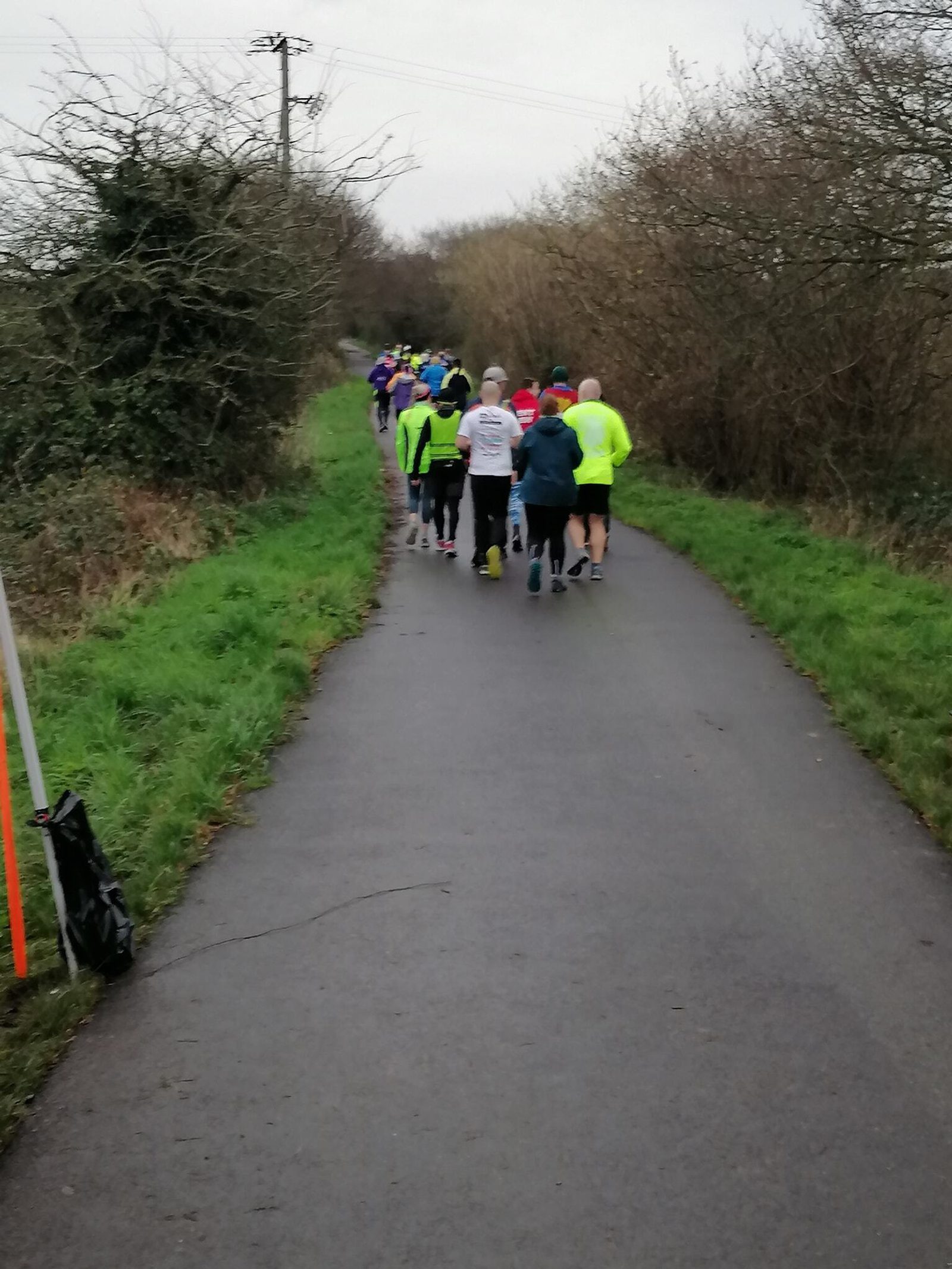 A group of people, wearing athletic clothing, running along a paved trail bordered by grass and leafless shrubs on a cloudy day. Some runners are in brightly colored outfits and appear to be participating in a group event, heading away from the camera.