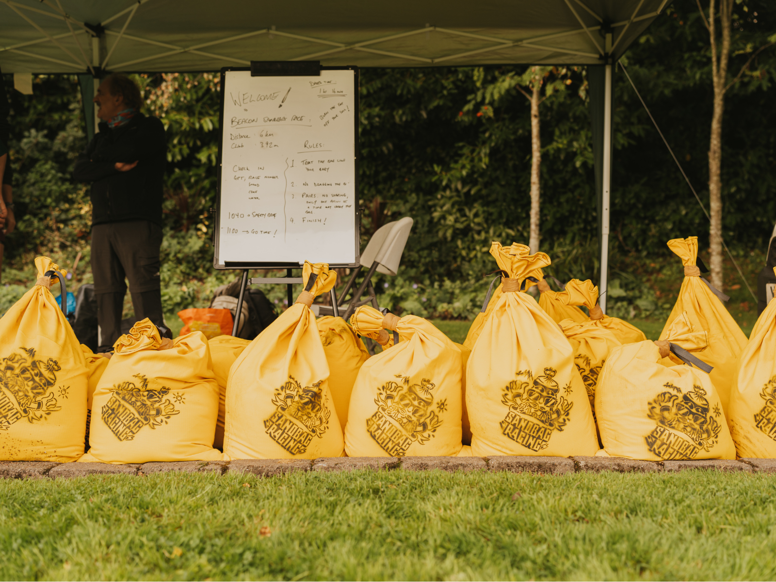 A row of yellow sacks arranged under a green canopy at an outdoor event. Behind the sacks, a whiteboard with written information is visible. Trees and grass are in the background, and a person stands to the left side of the image with their back turned.