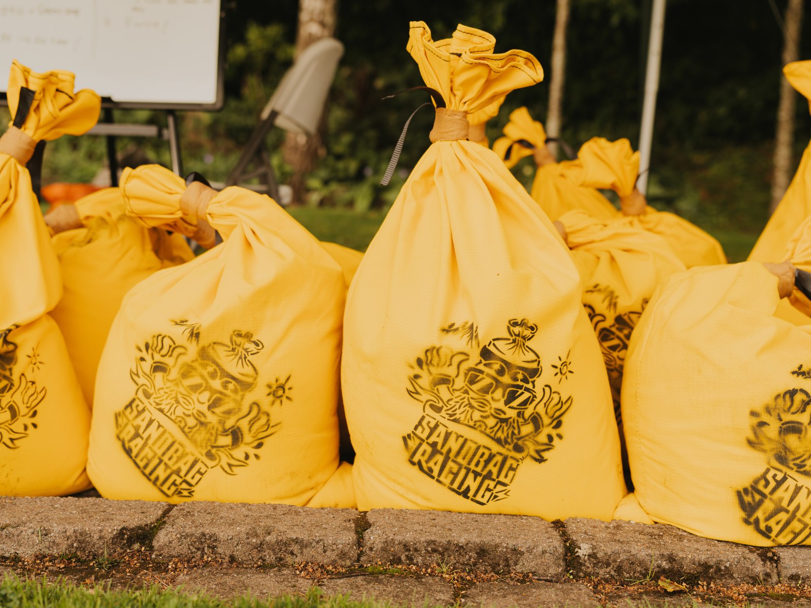 Several large, yellow sandbags lined up in a row on a paved surface. Each sandbag has a stylized logo with text that reads "SANDBAG RACING." Trees and greenery are visible in the background.