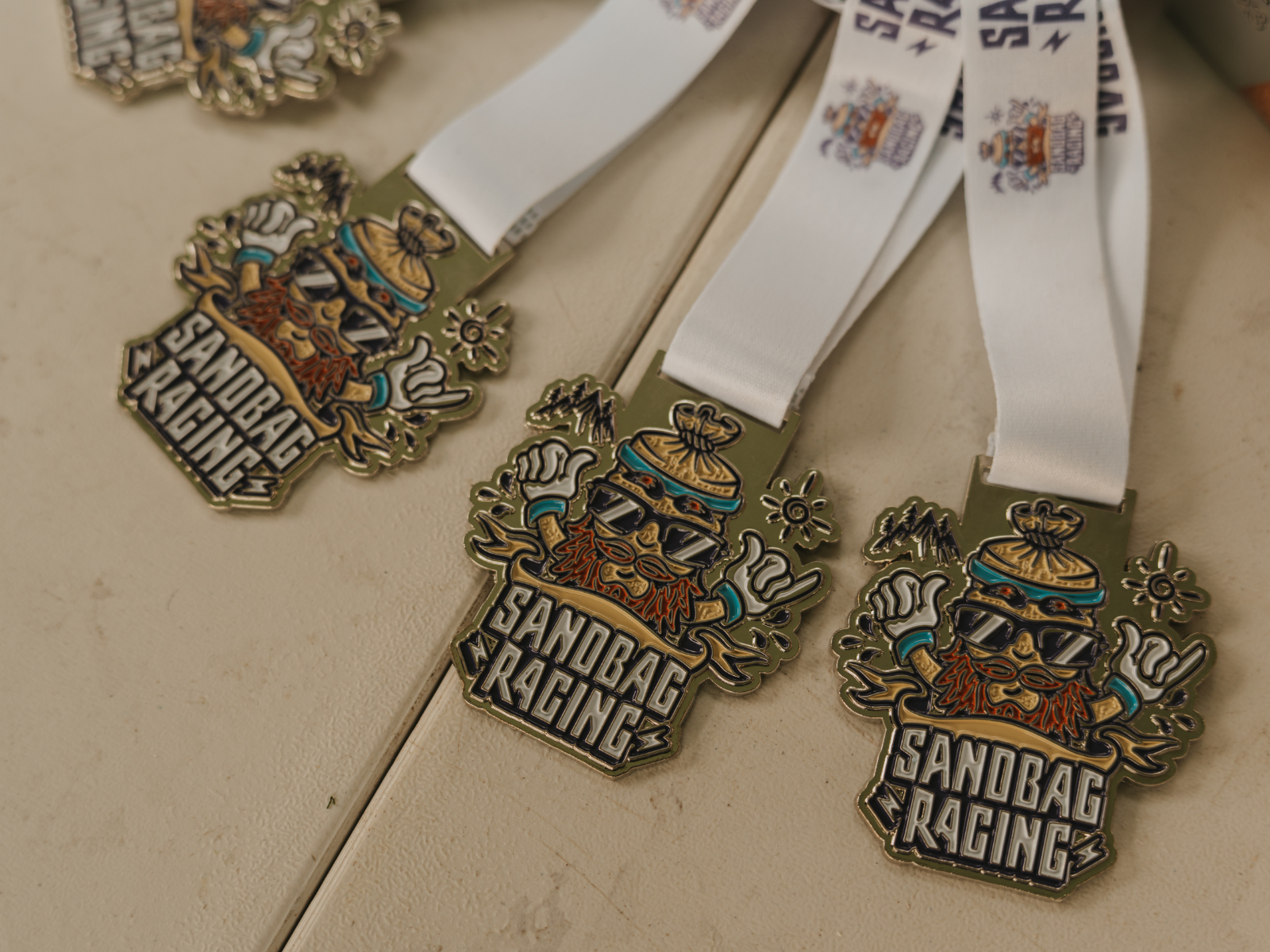 Close-up image of three "Sandbag Racing" medals on a table. Each medal has a colorful design featuring a bearded character with sunglasses and a helmet, attached to white ribbons with "Sandbag Racing" text and small graphics printed on them.