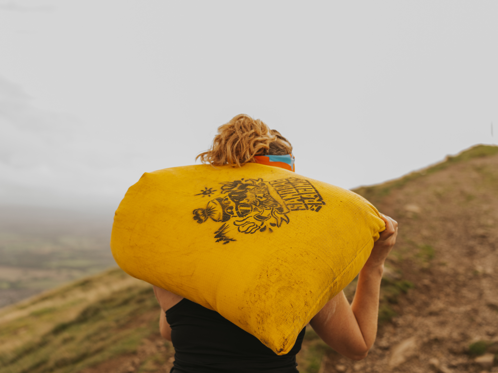 A person with blonde hair runs up a trail carrying a large yellow sandbag marked with an image and text on their shoulder. The background is an overcast sky with a misty, blurred landscape. The person appears to be participating in an outdoor fitness activity.