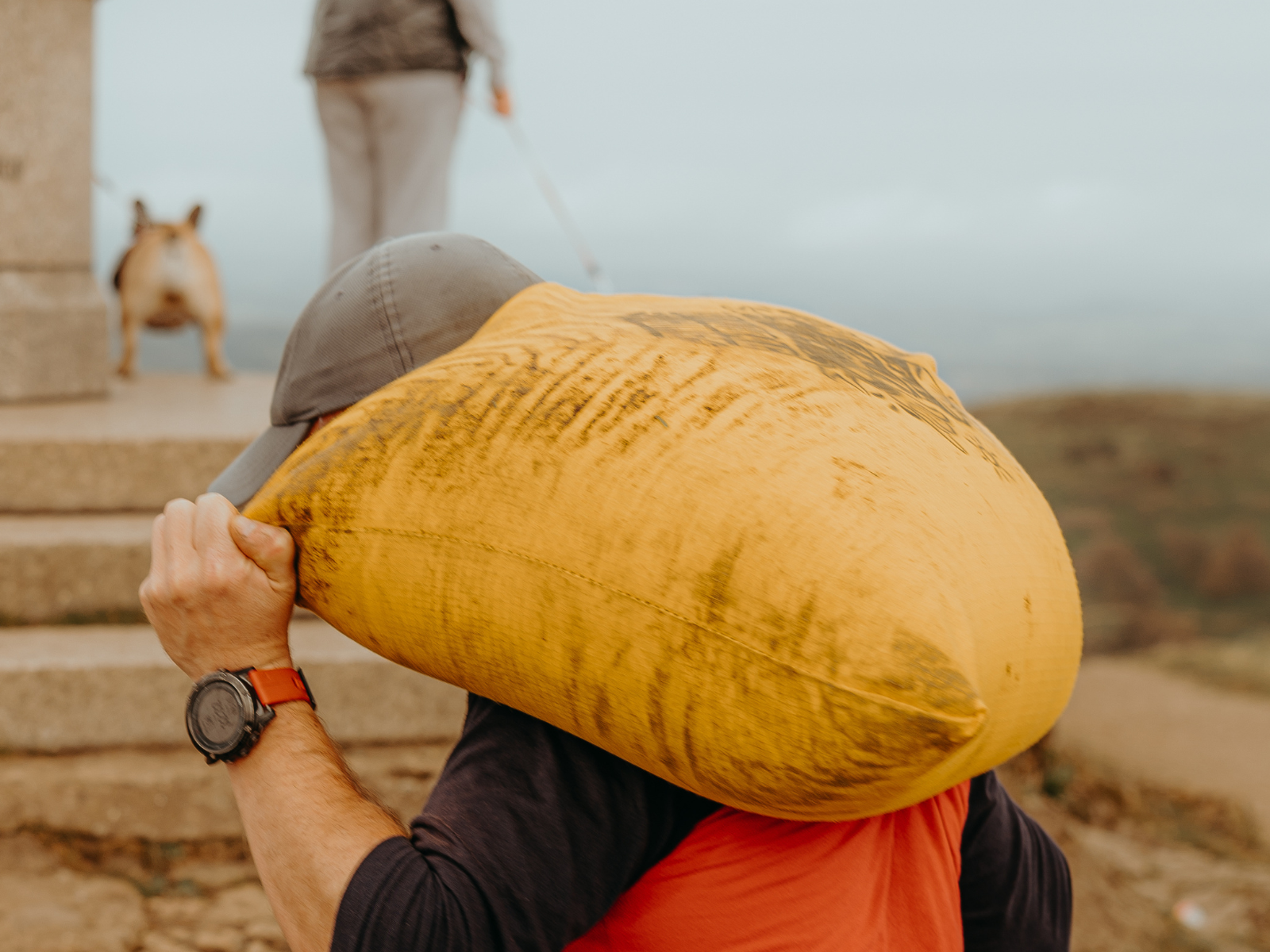 A person wearing a baseball cap carries a large, yellow sack over their shoulder while ascending stone steps. A small dog and another person with a hat are seen in the background. The setting appears to be outdoors on a cloudy day.