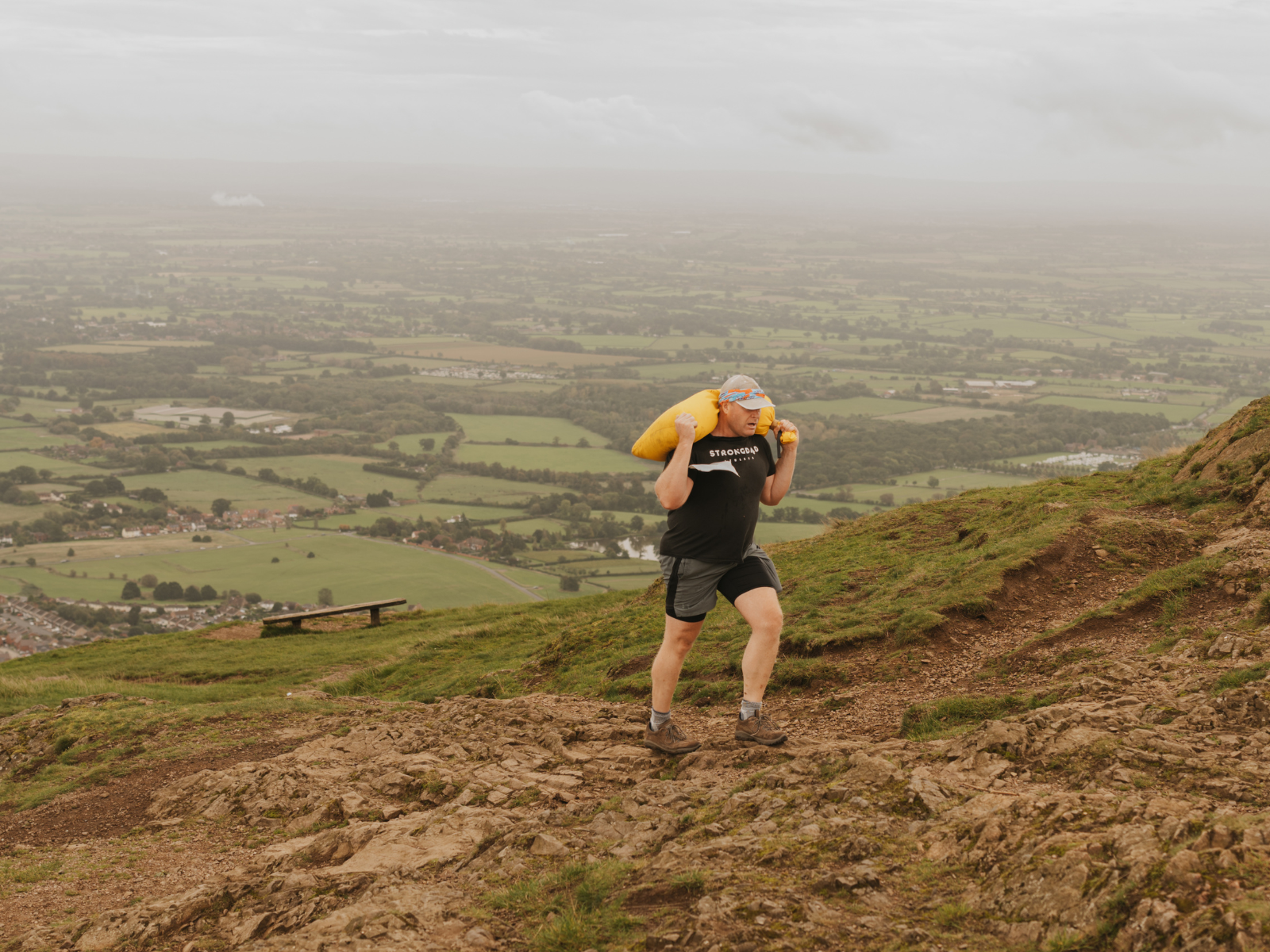 A person wearing a black T-shirt and shorts carries a large yellow sandbag on their shoulders while hiking up a rocky trail. The landscape in the background features green fields, distant trees, and a cloudy sky.