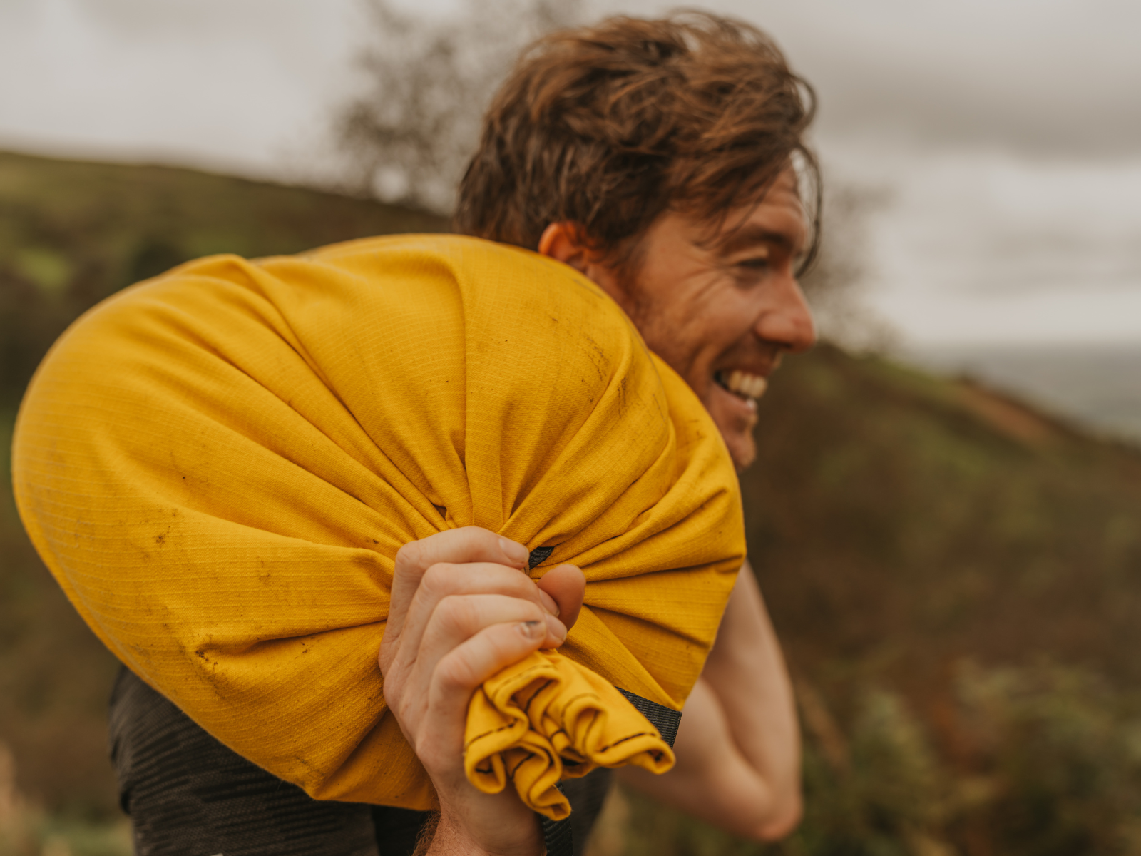 A smiling man with tousled hair carries a bright yellow bundle over his shoulder. The background is an overcast, blurred outdoor setting, possibly a hilly or mountainous area. The man appears to be in a casual, carefree mood.