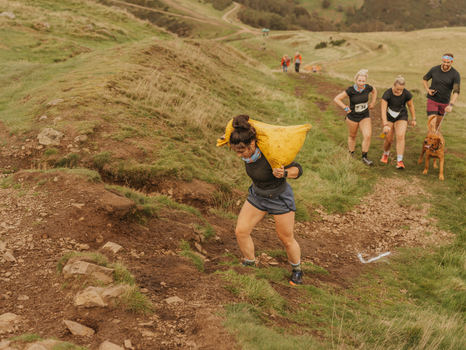 A woman in athletic gear carries a large yellow sack uphill on a rocky trail, followed closely by three other runners and a dog. The background shows a winding path through a grassy, hilly landscape on an overcast day.