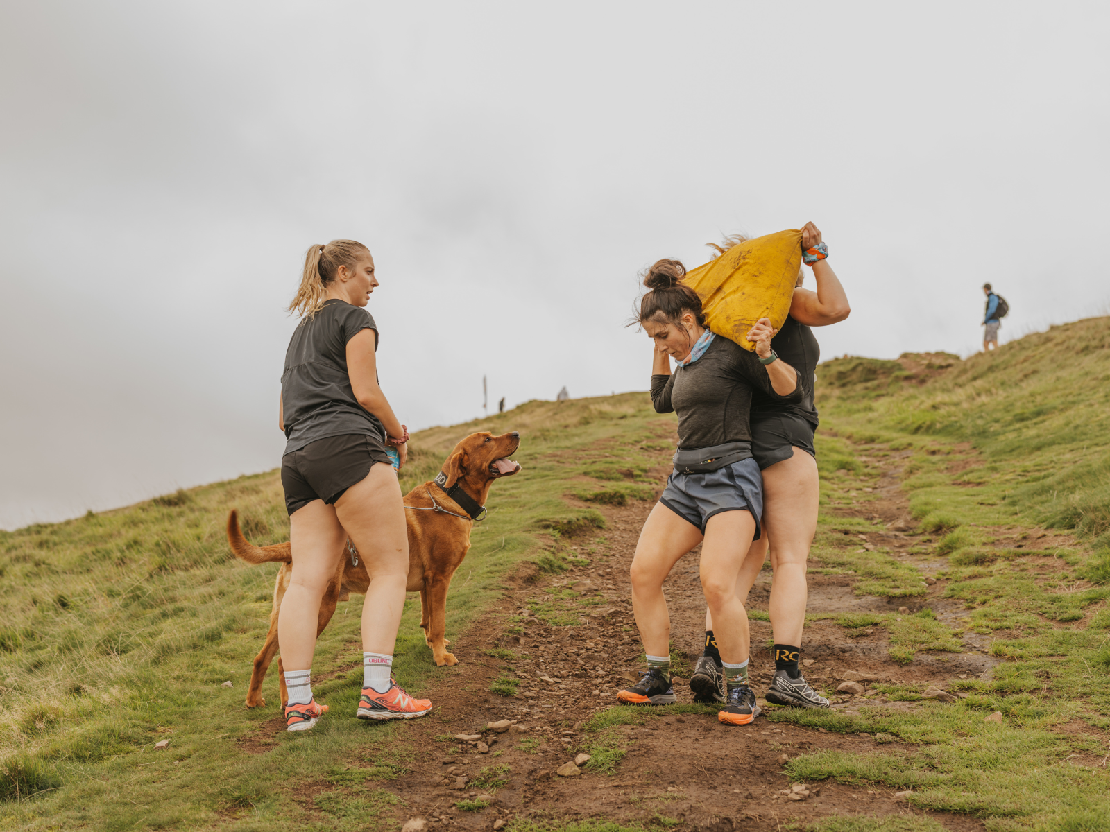 Three people are exercising on a grassy trail. One person carries a large yellow sandbag with another person assisting. A dog stands nearby, looking at them. The sky is overcast, and another person can be seen in the background.