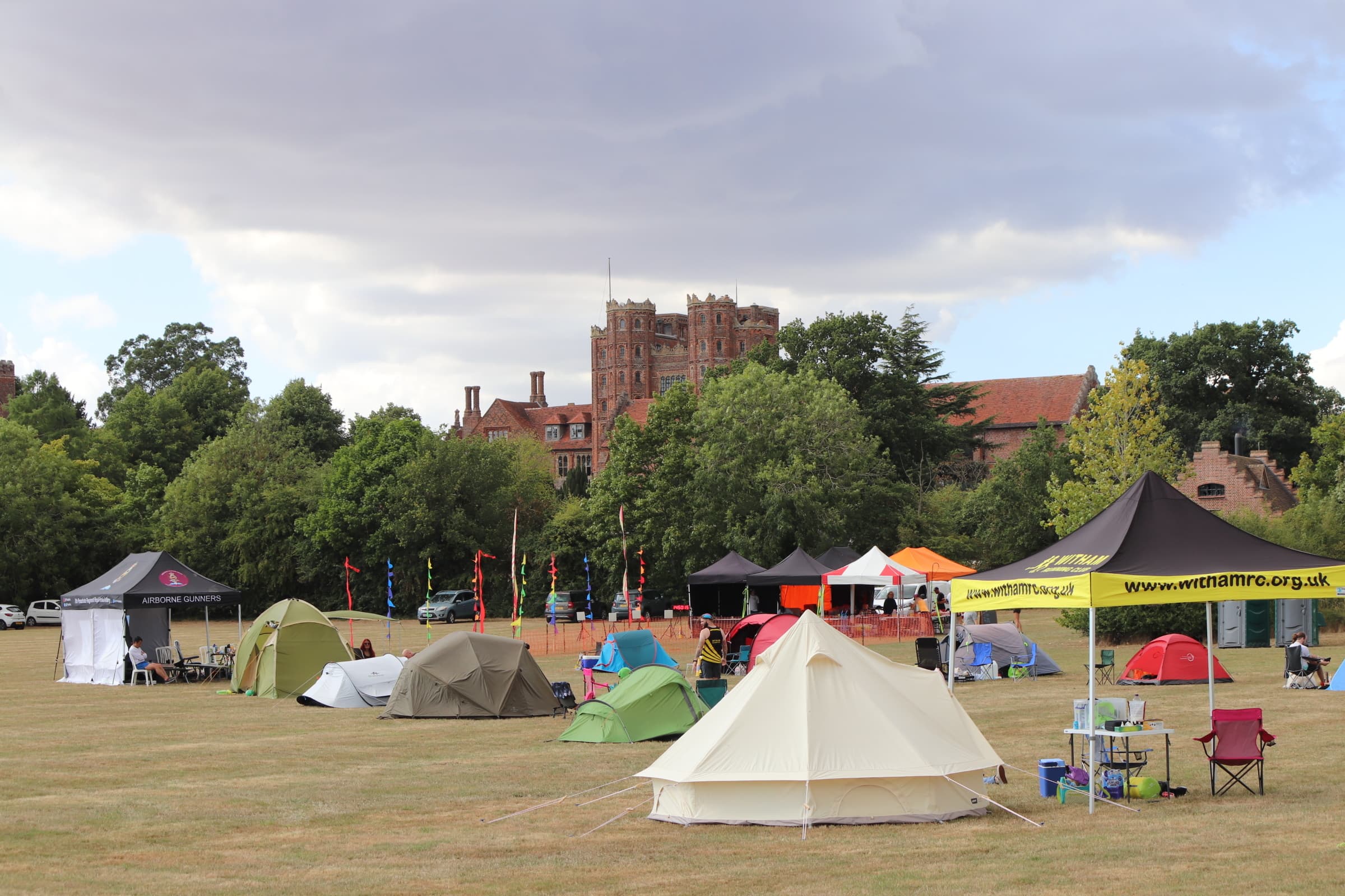 A large grassy field with numerous colorful tents and canopies set up. Various banners and flags are displayed. In the background, surrounded by trees, is an old, brick building with multiple stories and chimneys under a sky with clouds.