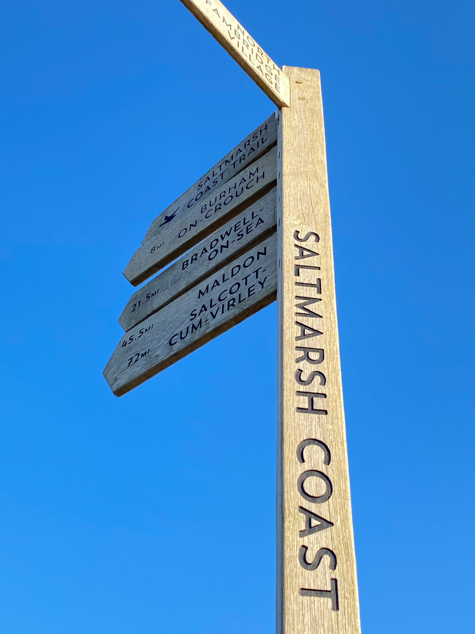 A tall wooden signpost against a clear blue sky. The post reads "SALT MARSH COAST" vertically. Several directional signs point to various destinations with distances, including "Bradwell on Sea 2.75M," "Maldon 8.5M," and "Salcott Cum Virley 7.25M.
