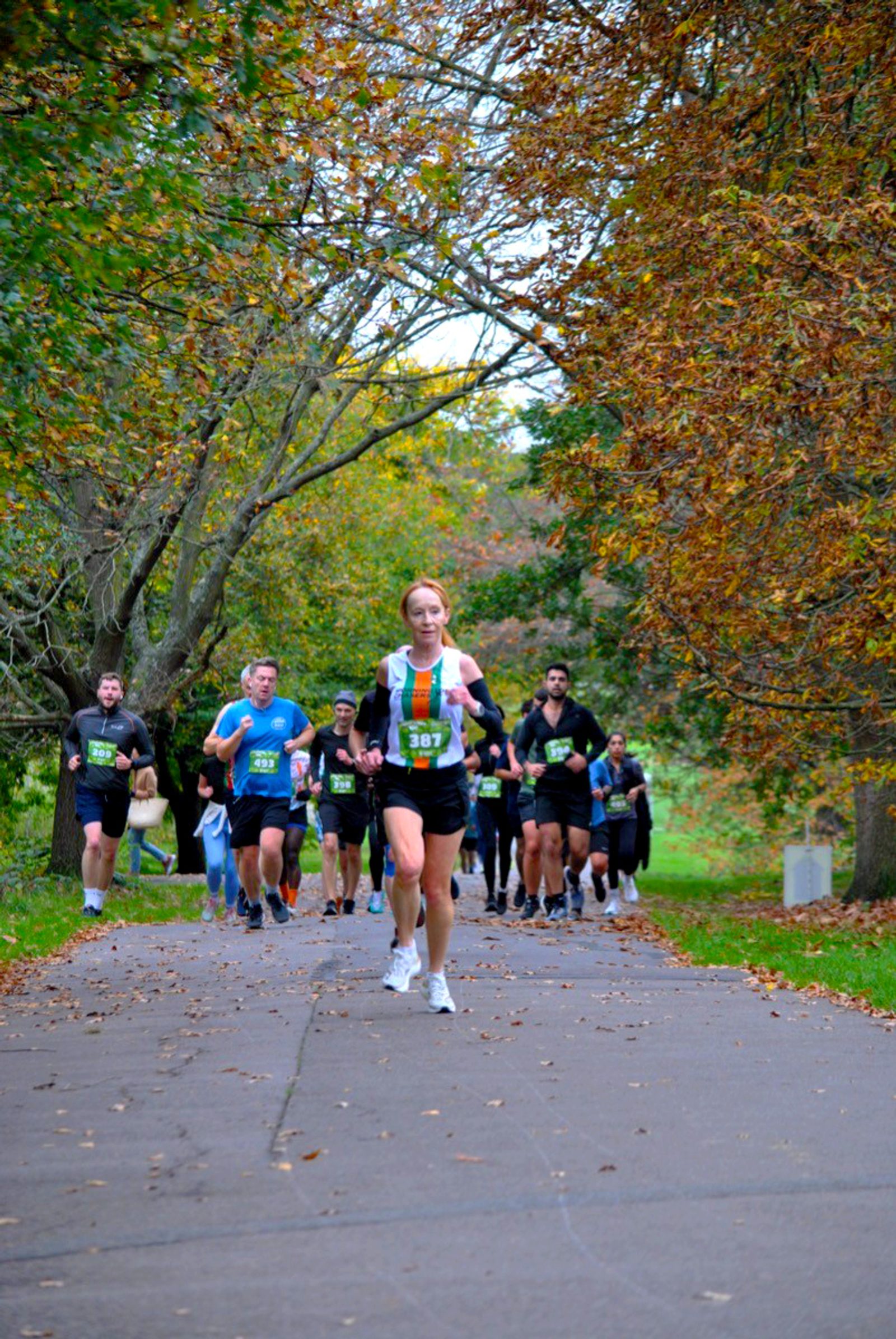 A group of runners participate in a race on a tree-lined path in autumn, with colorful leaves on the ground. One runner in a white and green vest leads the group.