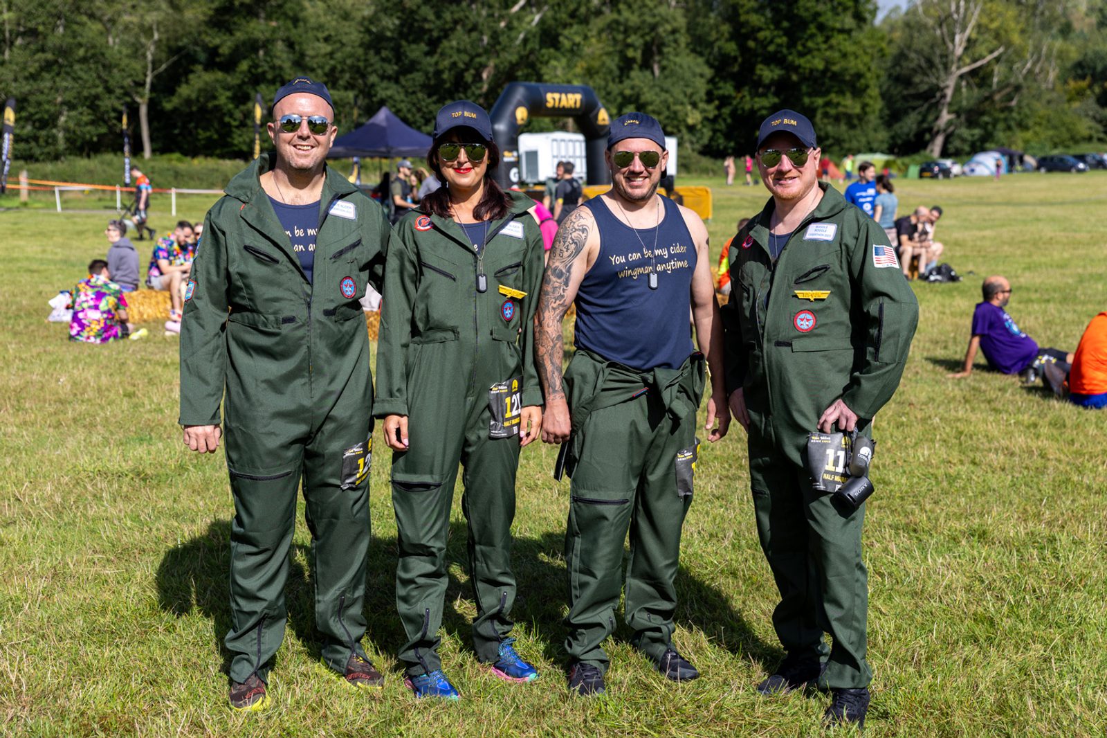 Four people dressed in green jumpsuits and navy caps are standing together on a grassy field. One person, second from the left, is wearing sunglasses. In the background, various people are seated on the grass, and a "START" inflatable archway is visible.