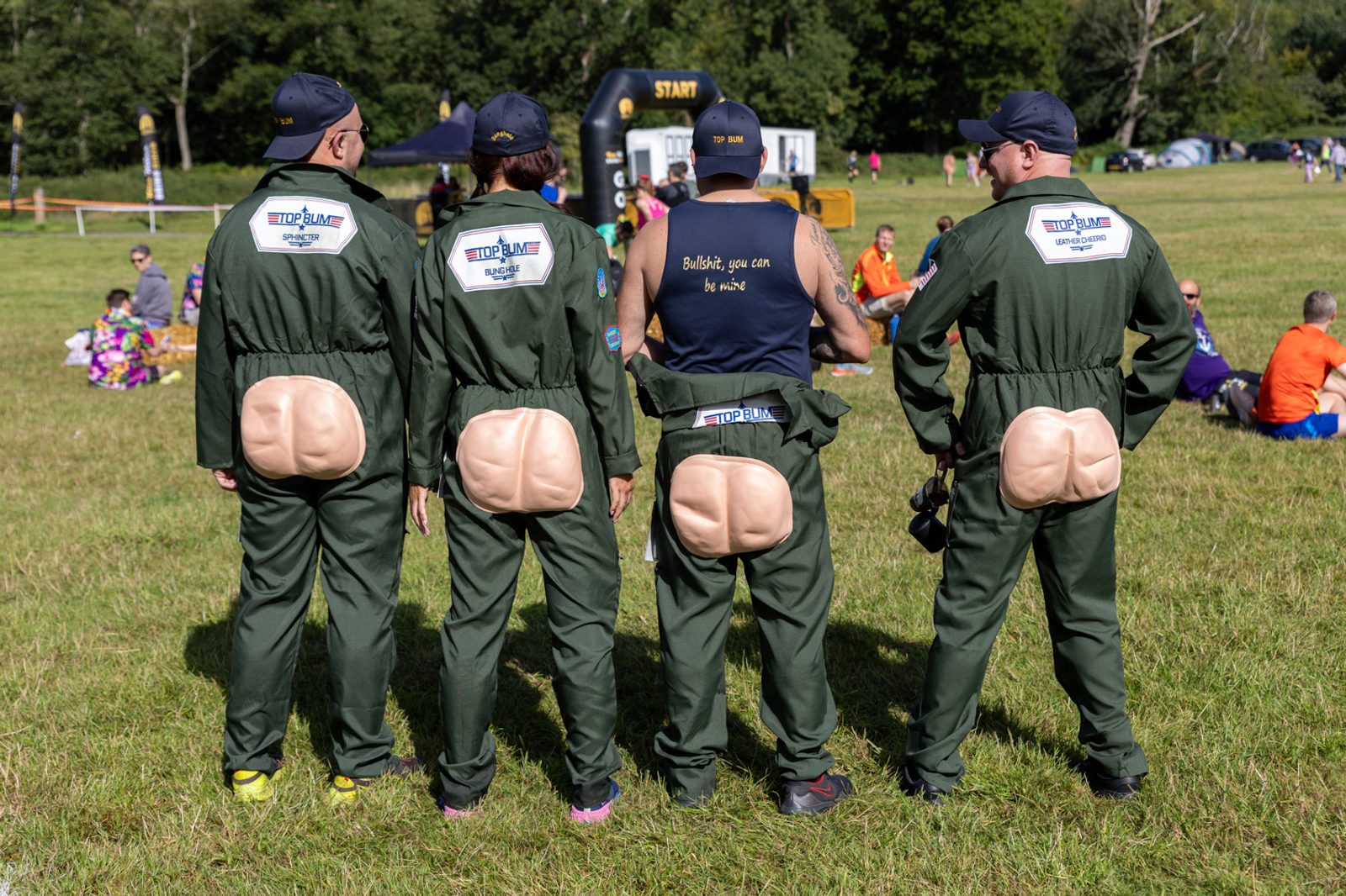 Four people stand with their backs to the camera, each wearing green jumpsuits with fake buttocks attached. They appear to be participating in a fun event at an outdoor field, with other groups and a starting line visible in the background.