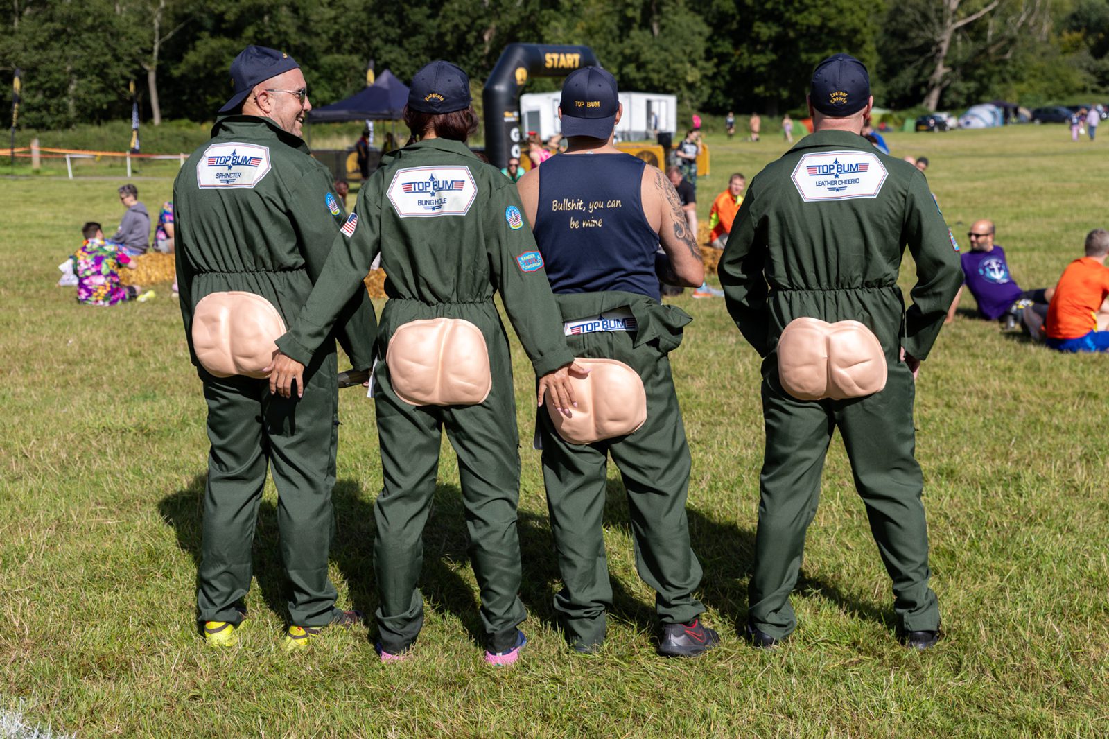 Four people standing outdoors, wearing matching green jumpsuits with fake buttocks attached to the back. They are facing away from the camera. Three of them are also wearing navy baseball caps. Other people are visible in the background, along with a finish line arch.