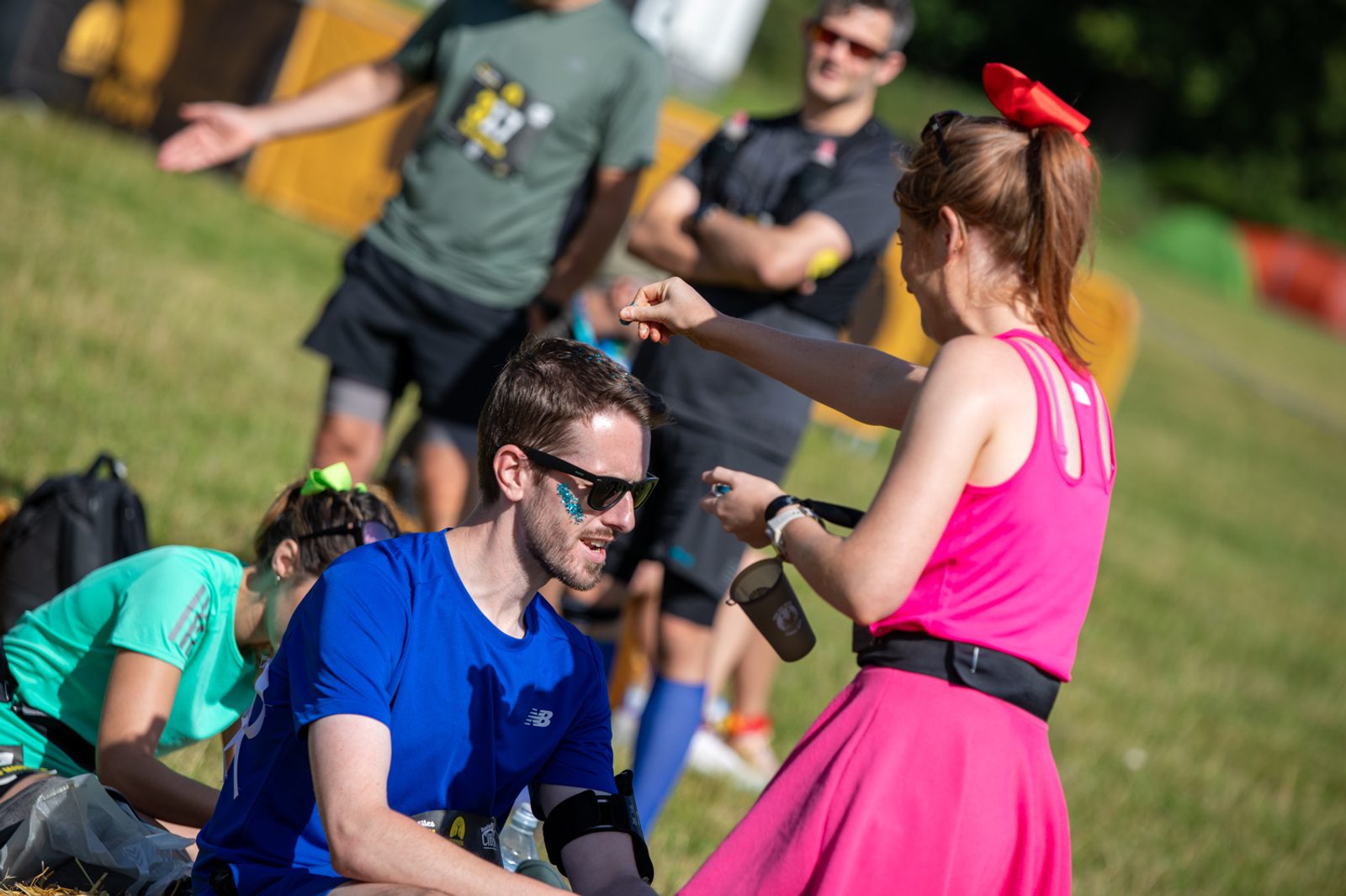A man in a blue shirt and sunglasses is kneeling on the grass, while a woman in a pink dress and red bow gestures towards him with a water bottle. In the background, two people stand and one person sits on the grass. It is a sunny day, and everyone appears relaxed.