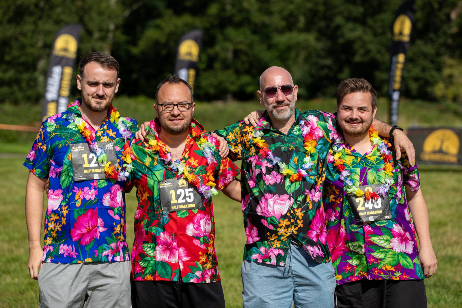Four men wearing colorful Hawaiian shirts and race bibs posing together outdoors. They are all smiling and standing on a grassy field, with trees and event banners in the background. Each man is adorned with a flower lei.