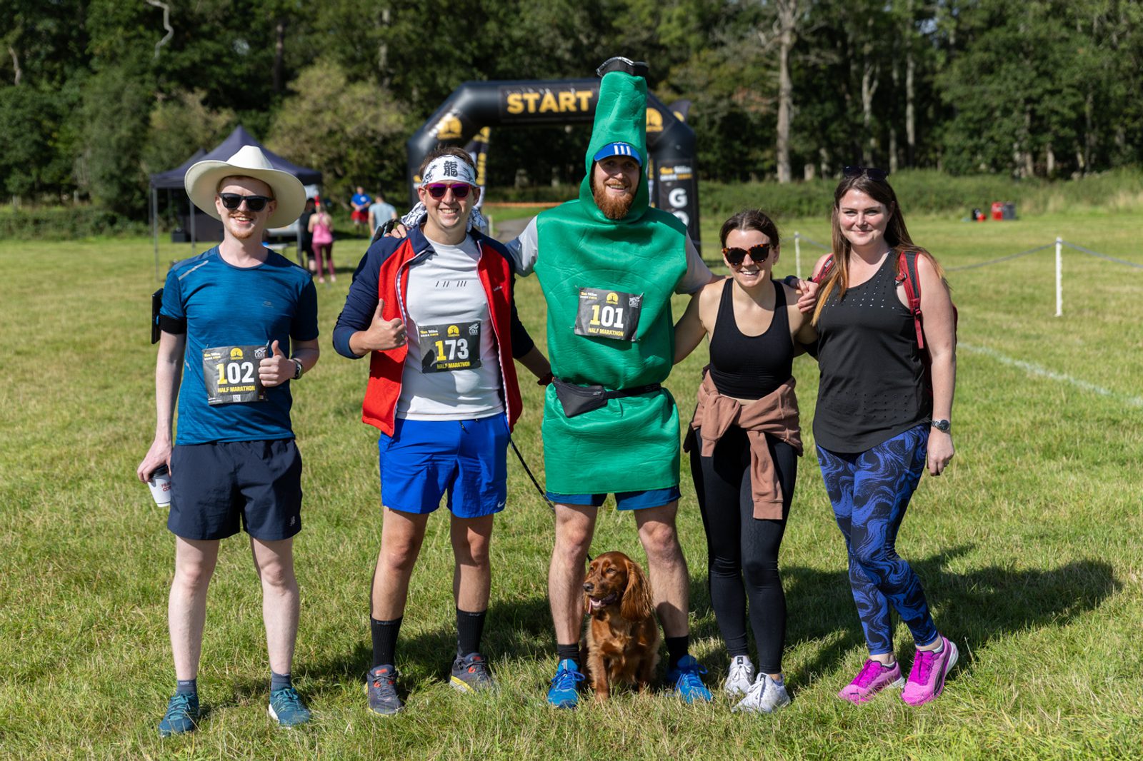Five people are posing for a group photo before a race on a grassy field with trees in the background. One person is dressed in a green costume, and a small dog sits at their feet. Four of them wear race bibs numbered 102, 173, and 101. An inflatable start line arch is visible behind them.
