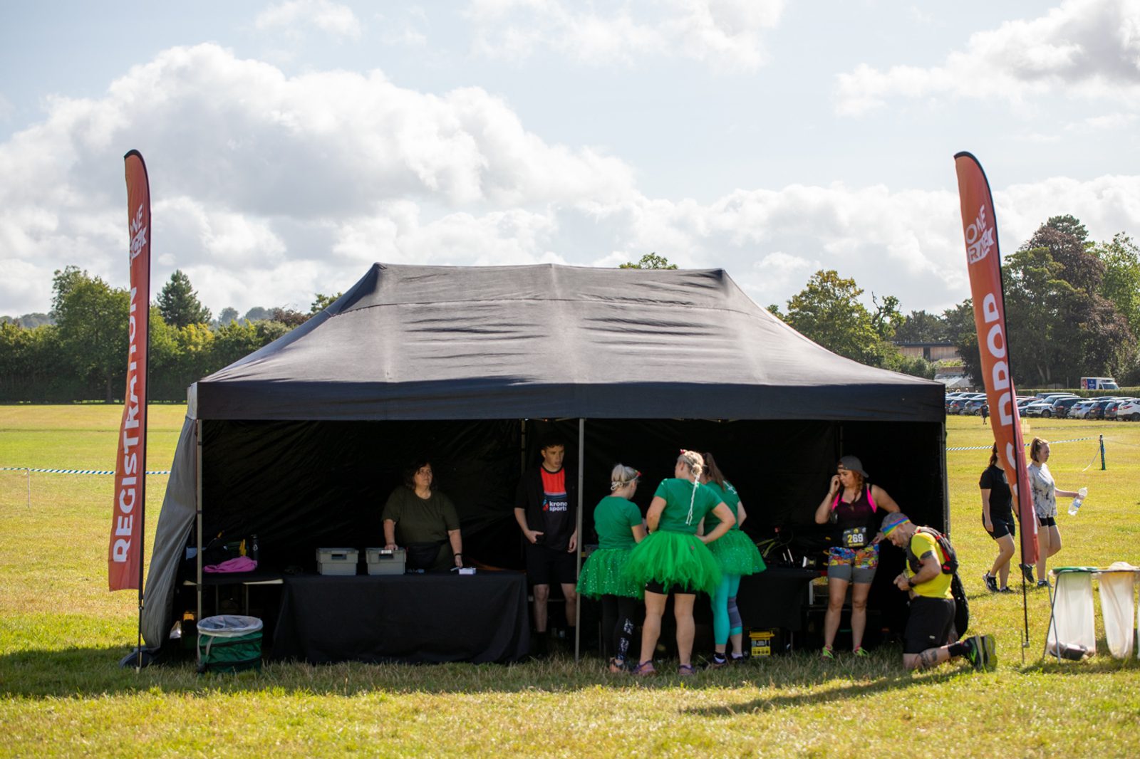 A black tent serves as the registration booth at an outdoor event. Several people are gathered around, with some wearing green costumes. Two tall orange banners labeled "REGISTER" and "INFO" are positioned on either side of the tent.