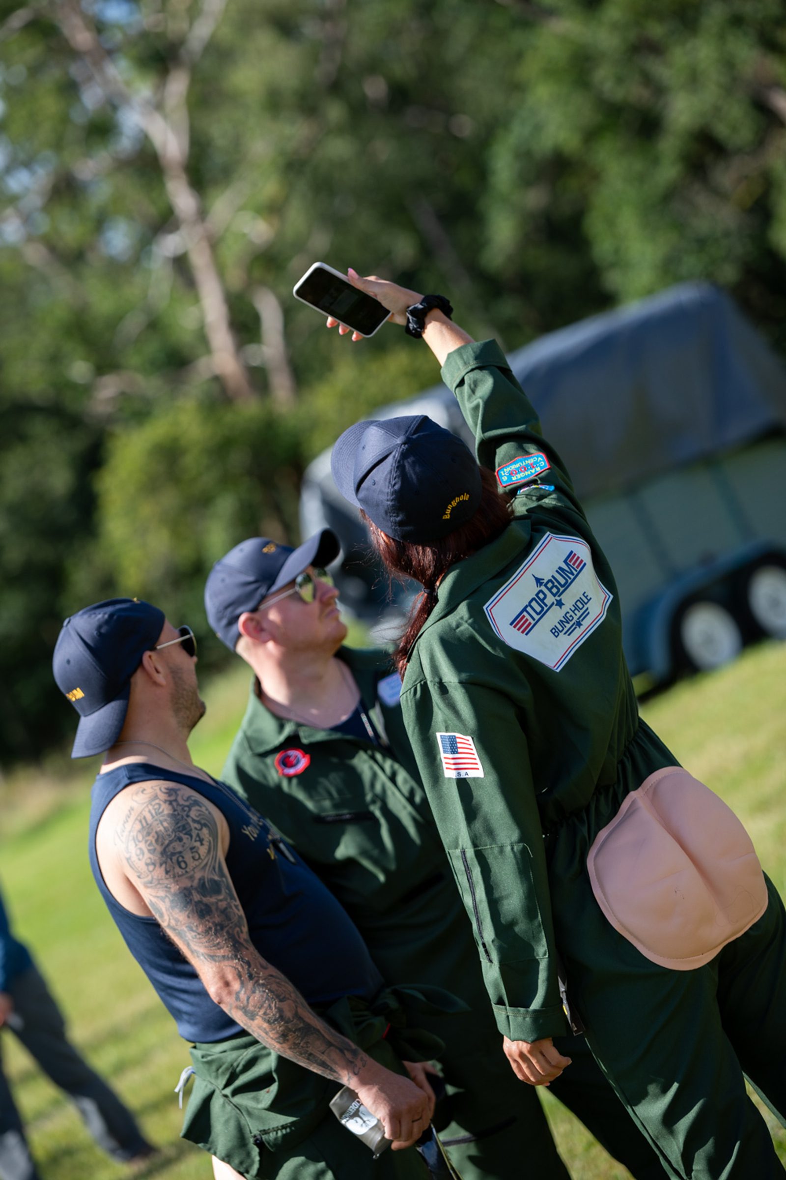 Three people in matching dark green jumpsuits and blue caps stand on a grassy field. Two men and one woman. The woman, with her back to the camera, holds up a smartphone as if taking a selfie. One man has sleeve tattoos, and a trailer is visible in the background.