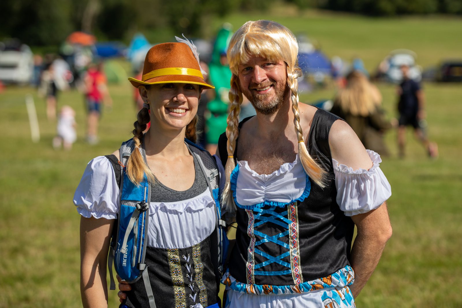 Two individuals stand outdoors at a daytime event, dressed in matching costumes with braided blonde wigs and traditional Bavarian-style attire. The person on the left wears a yellow hat and the person on the right smiles broadly. The background is a grassy area with people.
