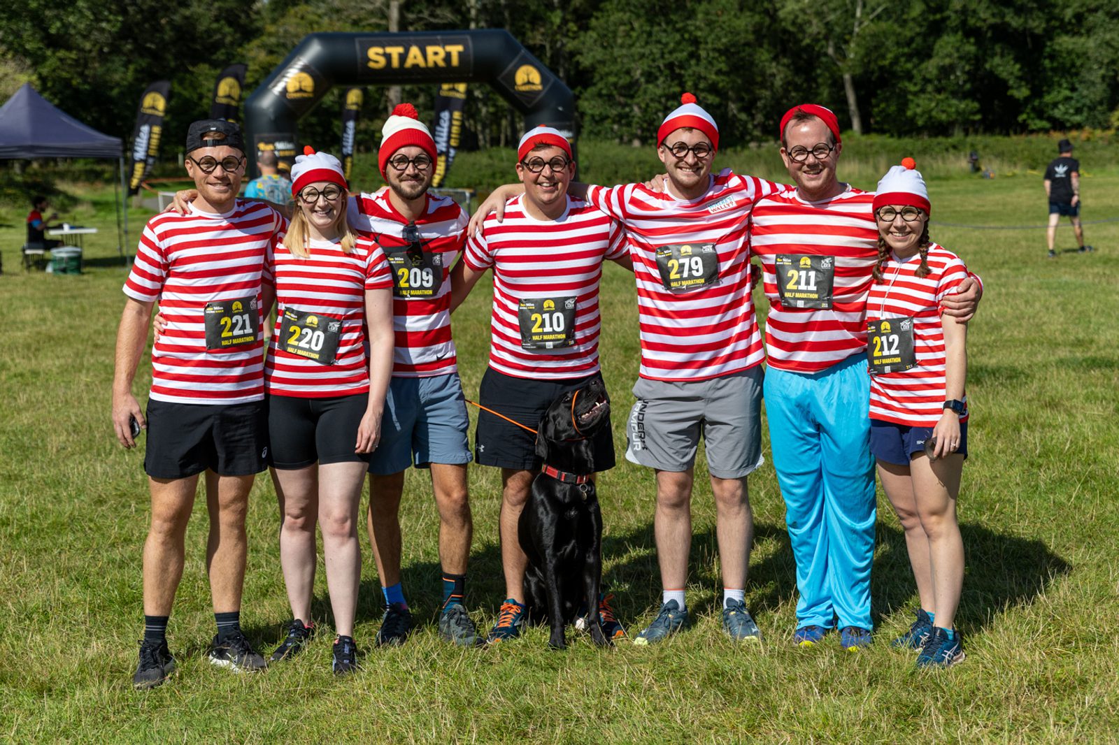 A group of seven people dressed in red and white striped shirts, some with hats, and one with a dog, stands in a grassy field in front of an inflatable start line. They appear ready to participate in a race or a themed event, smiling and posing for a photo.