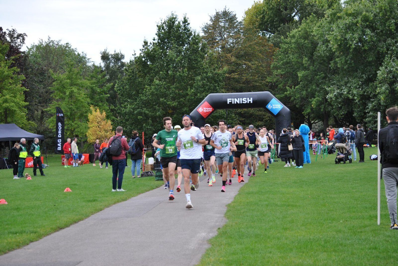Runners sprint toward the finish line during a race in a park, surrounded by spectators, trees, and event tents. The "FINISH" arch is visible in the background.