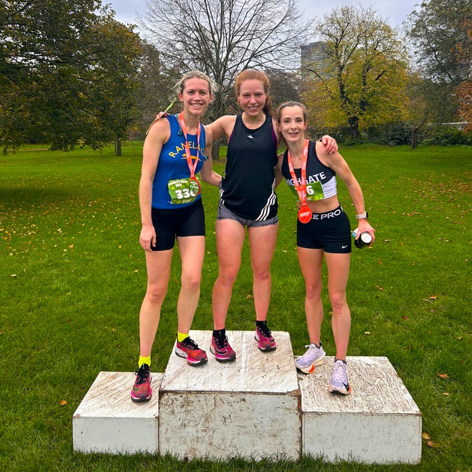 Three smiling female runners with medals stand on a winners' podium outdoors in a park, dressed in athletic gear, with autumn trees and grass in the background.