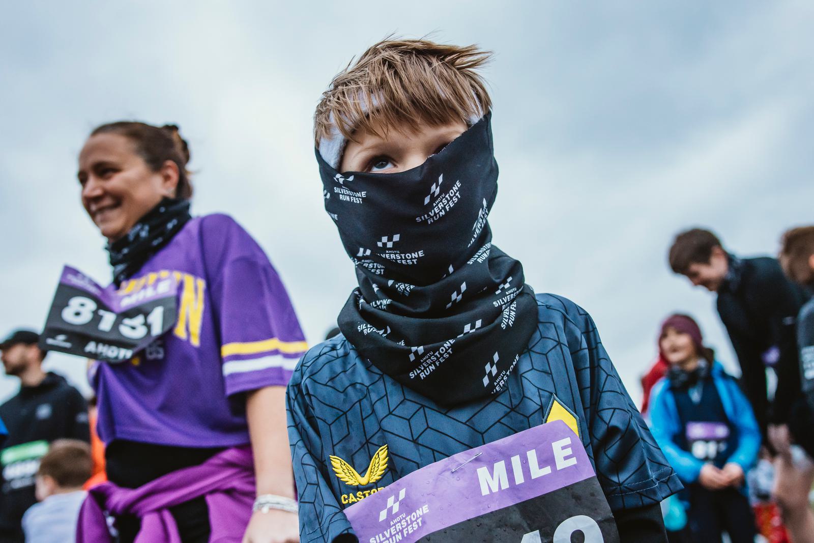 A young participant wearing a patterned face covering stands at an outdoor event. He wears a numbered bib and a dark shirt with a logo. Other people in athletic gear are in the background under a cloudy sky.