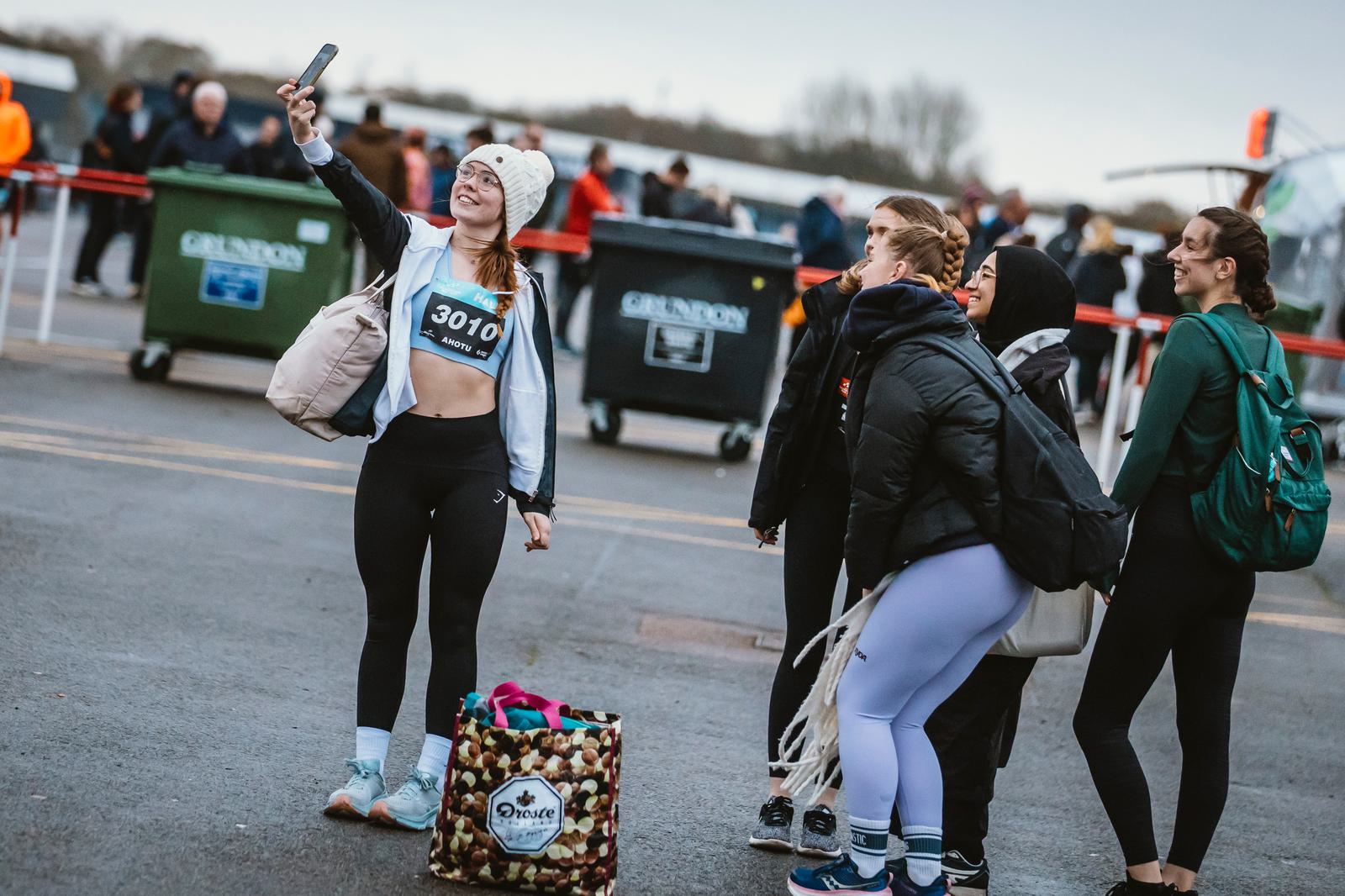 A group of five people in athletic wear smile as one takes a selfie. They stand in a parking area with bins in the background, dressed warmly for cool weather, with bags at their feet. The person with the phone wears a beanie and has race number 3010.
