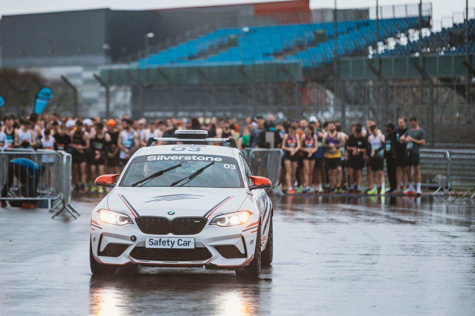 A BMW safety car with "Silverstone" written on it leads a group of runners at an outdoor race event. The ground is wet, and the scene includes a stadium in the background.
